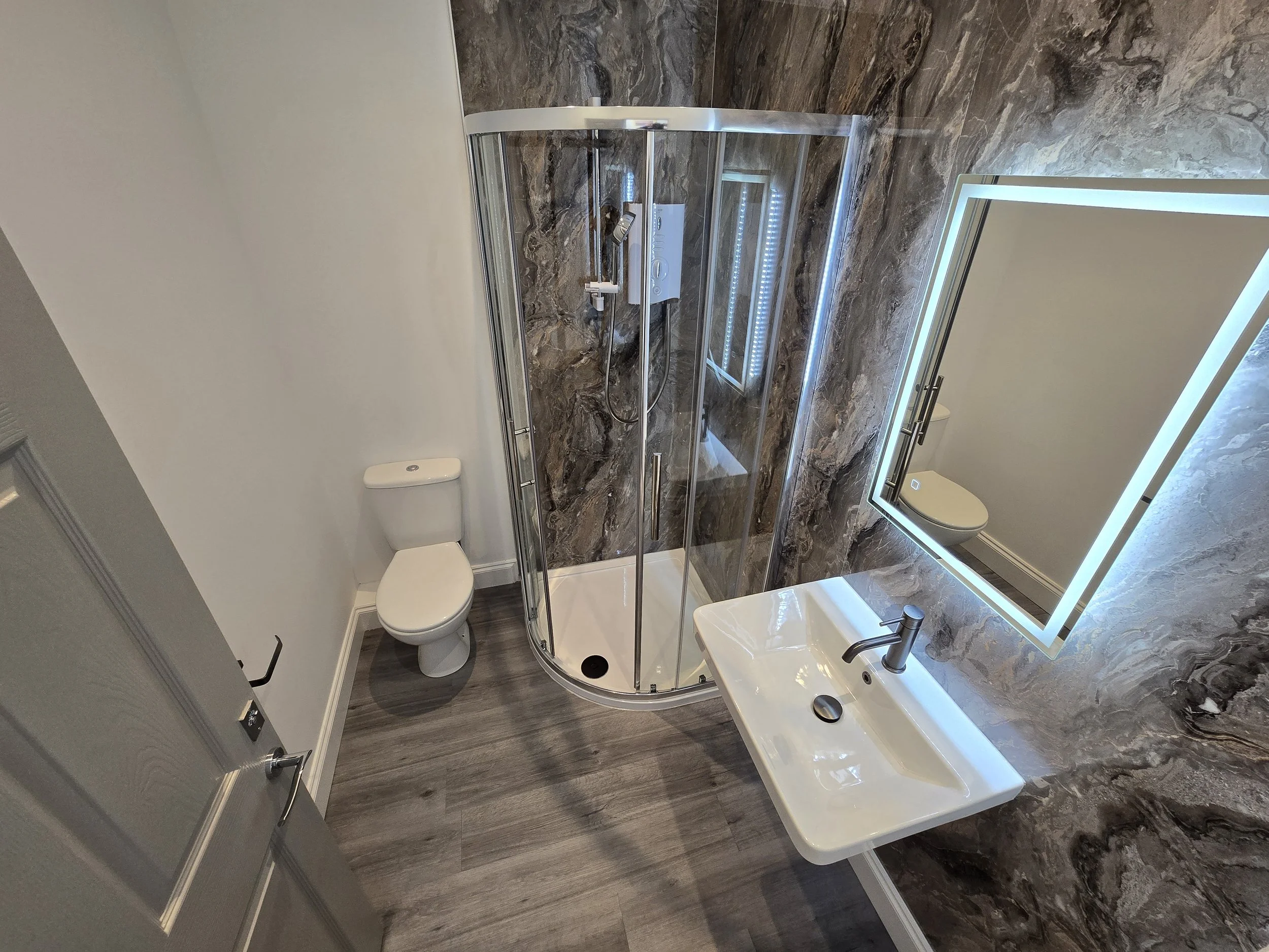 A modern bathroom with a white toilet, a white sink with a metallic faucet, a large rectangular illuminated mirror, a rounded corner glass shower with marble walls, and gray wooden-style flooring.