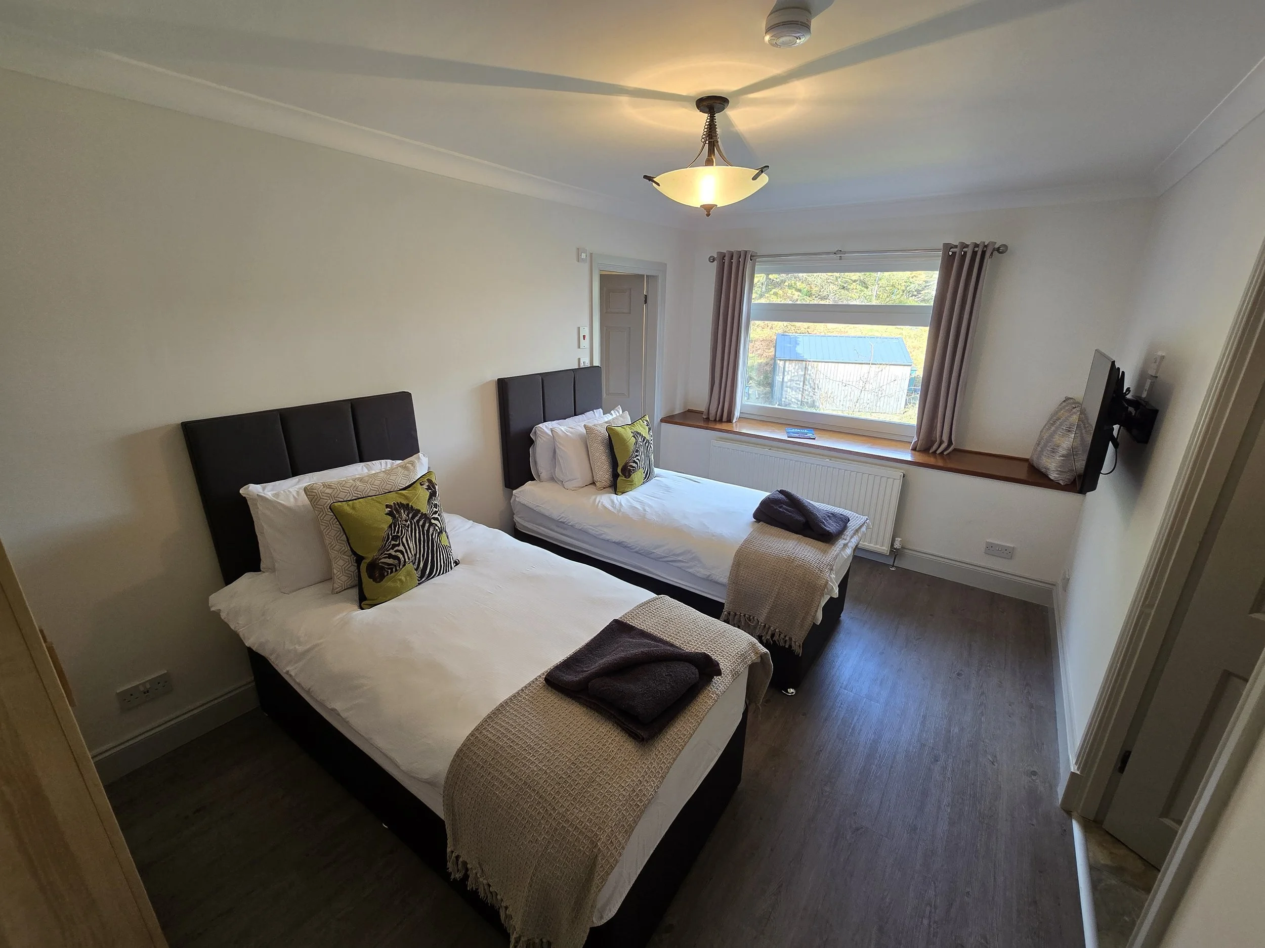A bedroom with two single beds, each with black headboards, white bedding, and zebra-themed throw pillows. The beds have folded towels at the foot. There is a window with beige curtains, and a flat-screen TV mounted on the wall. The room has a ceiling light, wooden flooring, and a small ledge underneath the window with some items on it.
