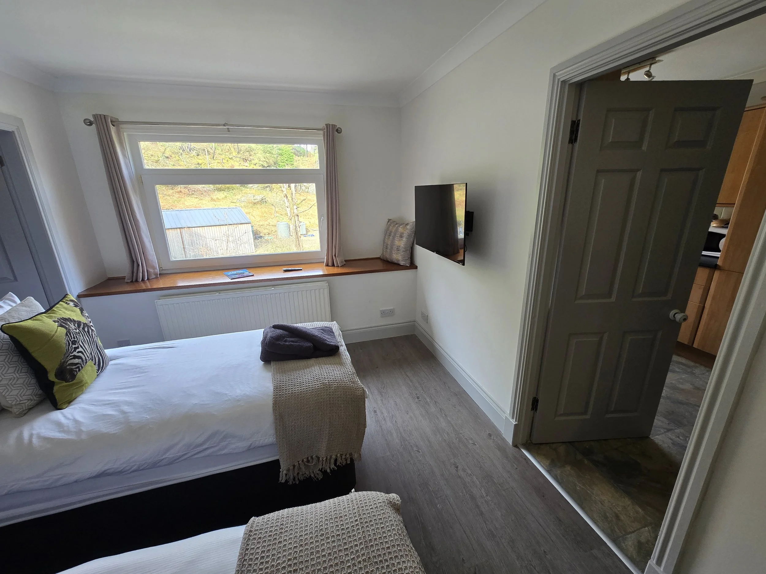 A cozy bedroom with a large window, bed with zebra and animal print pillows, a beige knit blanket, a wall-mounted flat-screen TV, and an open door leading to a kitchen area.