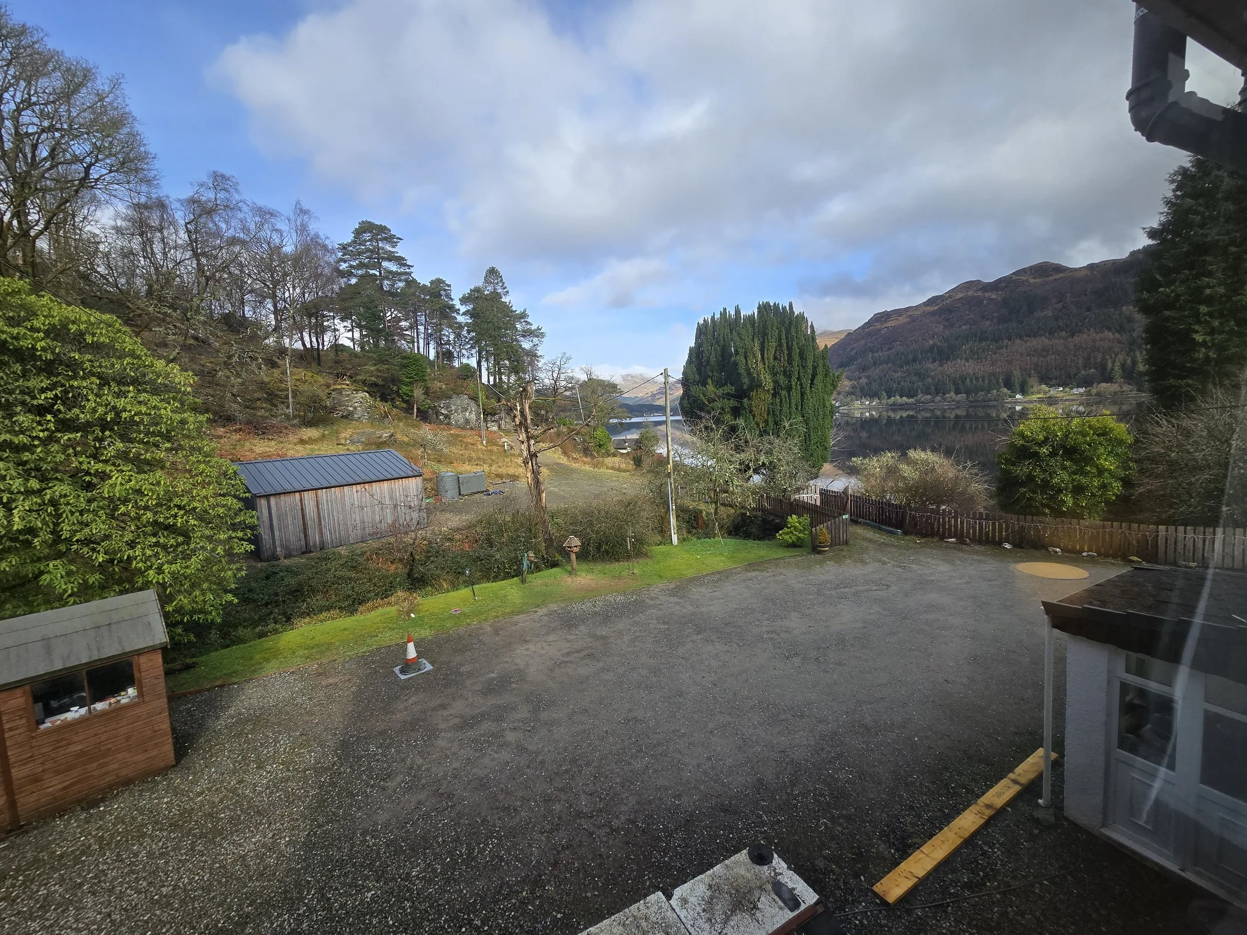 Landscape view of a backyard with trees, a gravel area, and a lake with mountains in the background. Part of a house is visible in the foreground.