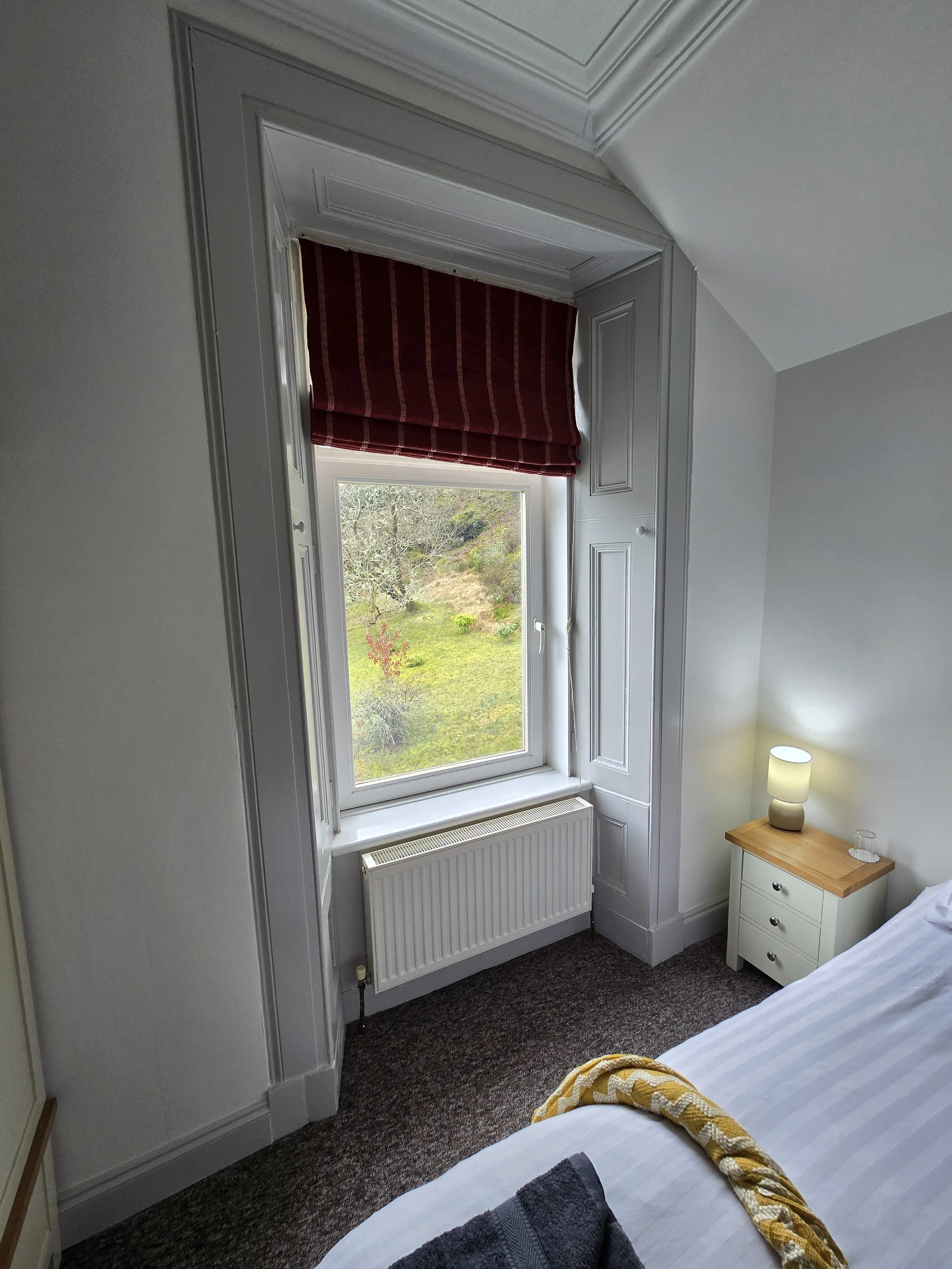 A bedroom window with white wooden trim and a red-striped window shade. Outside view of trees and grass. Part of a bed with a yellow patterned blanket and a nightstand with a lamp.