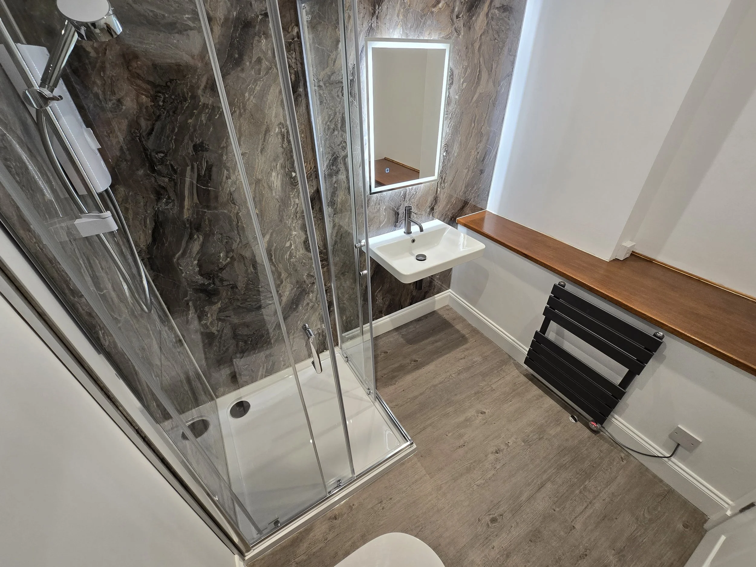 Small bathroom with a corner shower, white sink, mirror, and wood shelf. The floor is wood-look laminate, and there's a black heater on the wall.