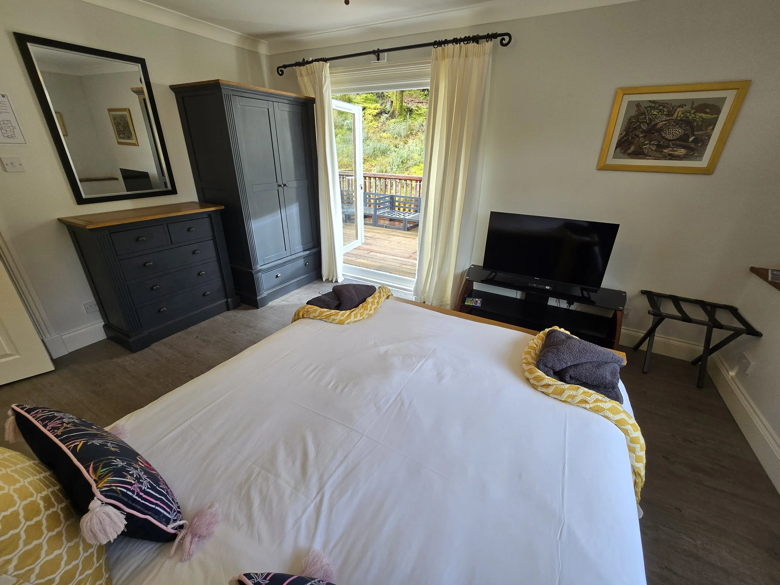 A cozy bedroom with a bed with white sheets and decorative pillows, dark furniture including a dresser, wardrobe, and TV stand, and a balcony with a view of greenery outside.