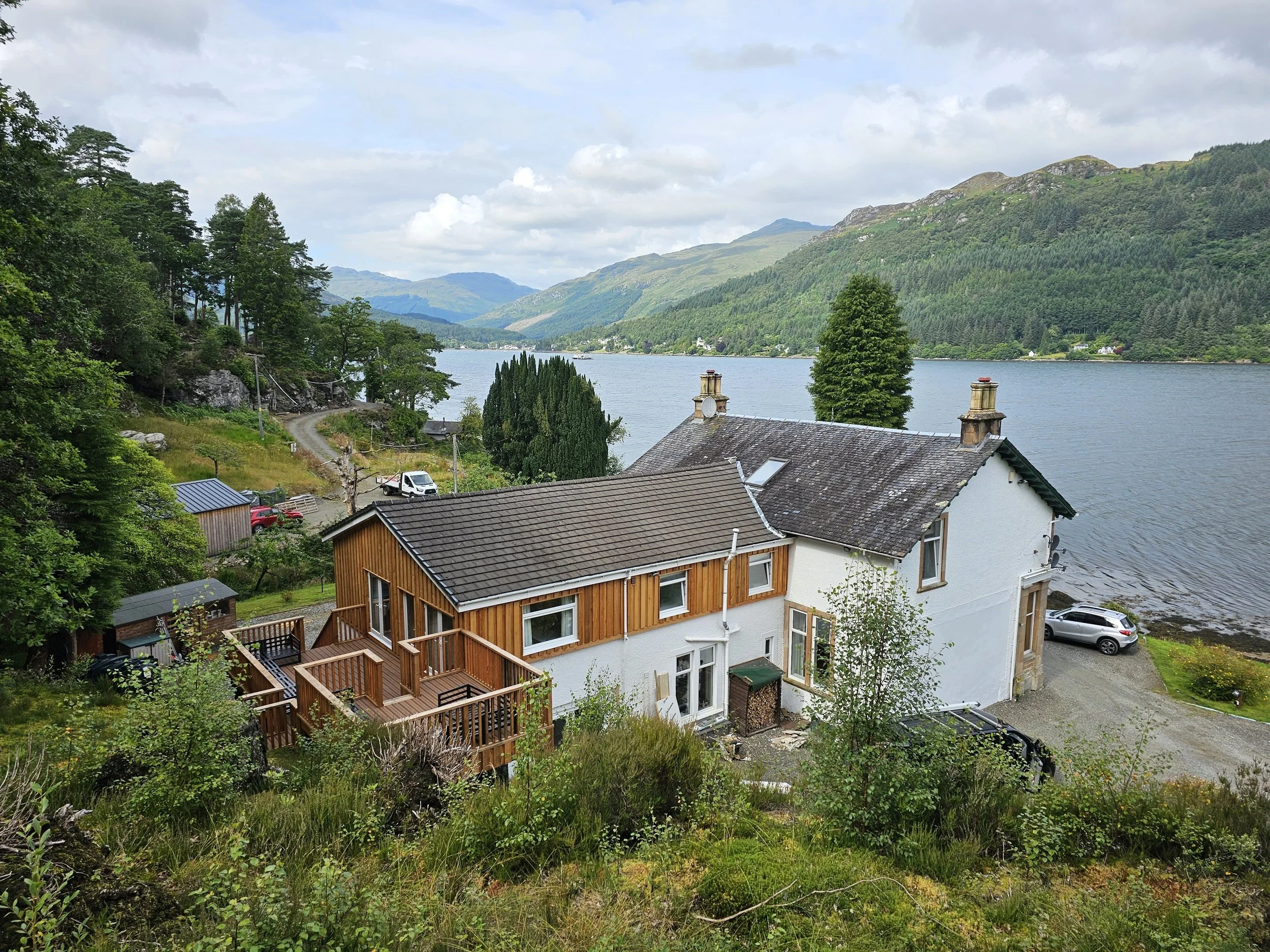 A house by a large loch with green mountains in the background, surrounded by trees and vegetation.