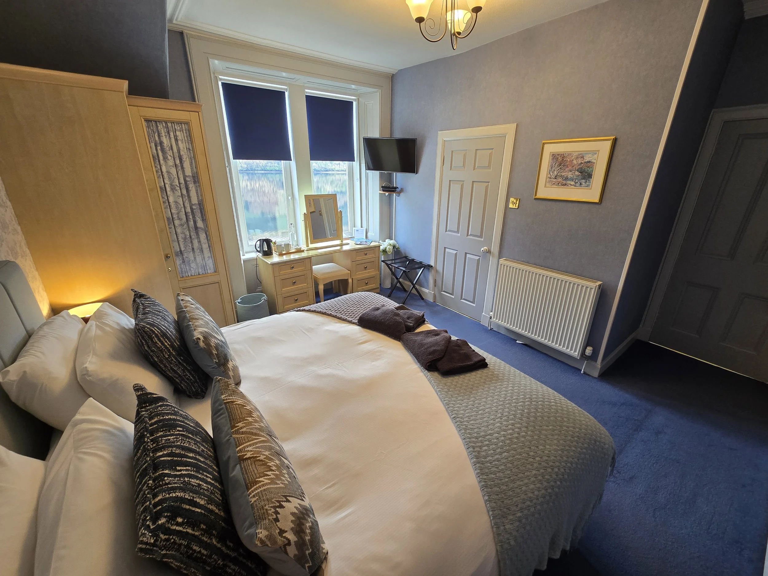 A cozy hotel bedroom with a double bed, decorative pillows, a nightstand, a wardrobe, a window with blue Roman blinds, a dressing table with a mirror, a small flat-screen TV, and a radiator. The room has soft blue walls and a ceiling light fixture.