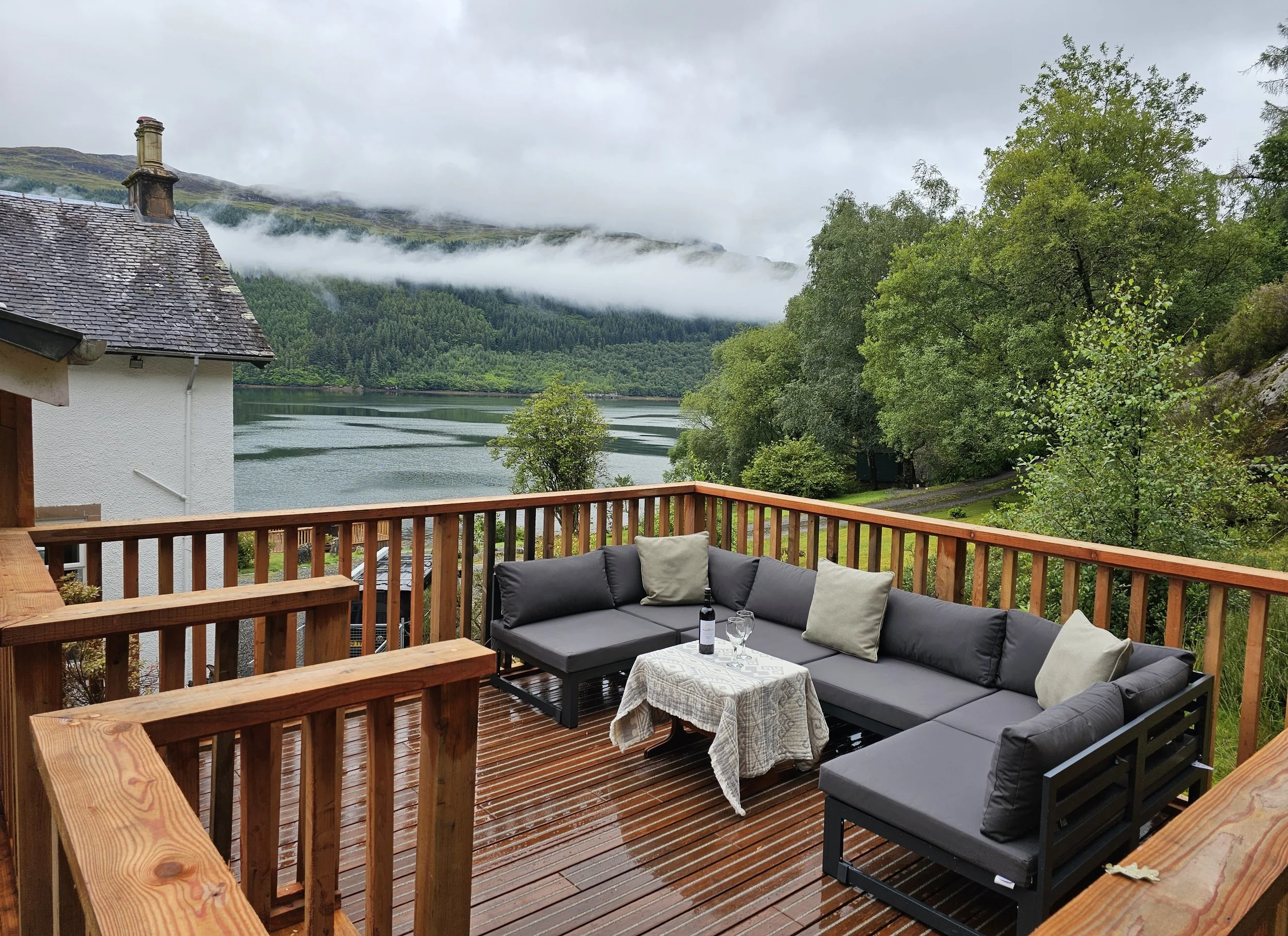 A wooden deck with outdoor seating, including a dark gray sectional sofa with pillows, overlooking a lake surrounded by green trees and mountains with low clouds in the background.