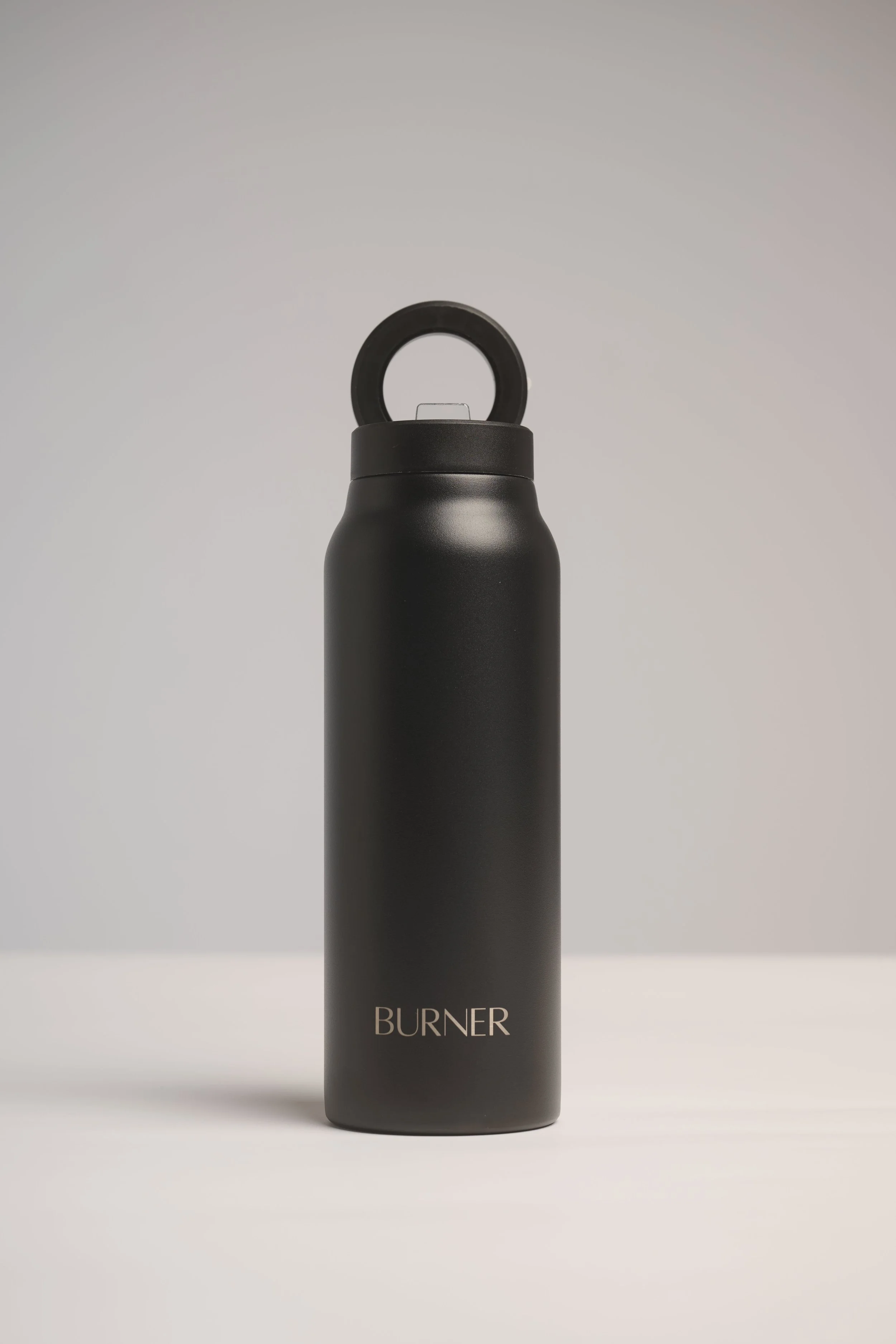 A black bottle with a circular handle on top, labeled 'BURNER' in gold text, standing against a plain, light background.
