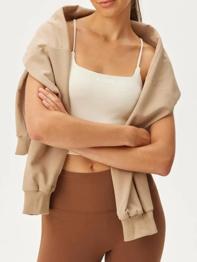 A woman with crossed arms wearing a white crop top with spaghetti straps, a beige hoodie draped over her shoulders, and brown high-waisted leggings.