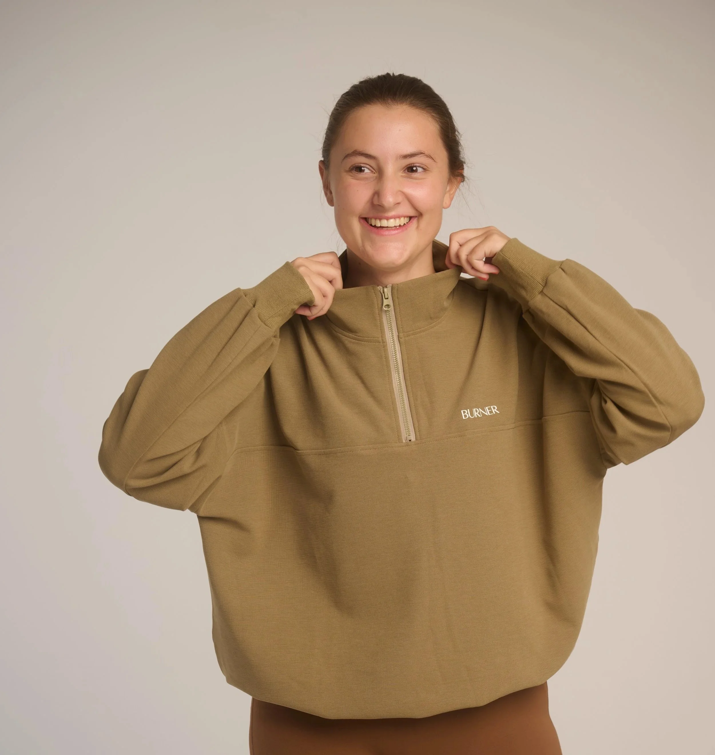 A young woman with brown hair tied back, wearing a tan hoodie and smiling, standing against a neutral background.