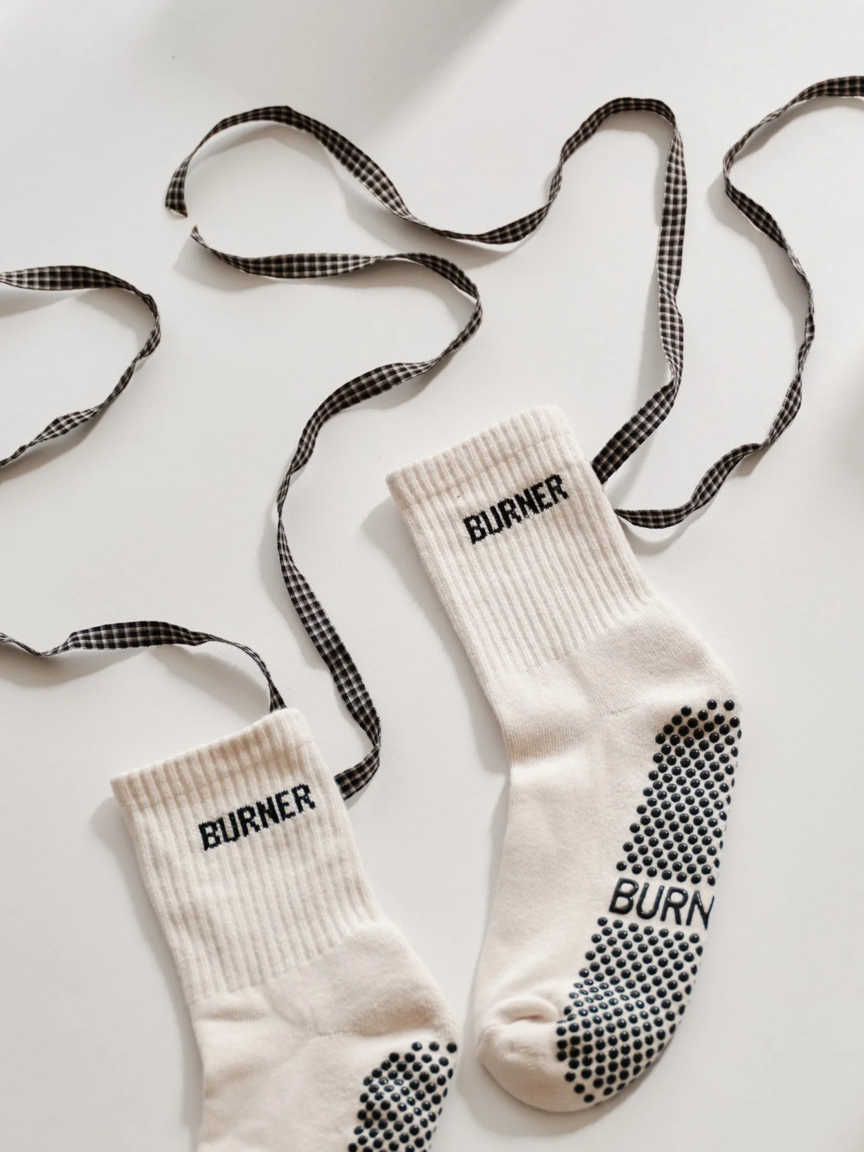 Pair of white socks with black text "BURNER" and black dotted soles, connected by a black and white checkered shoelace.
