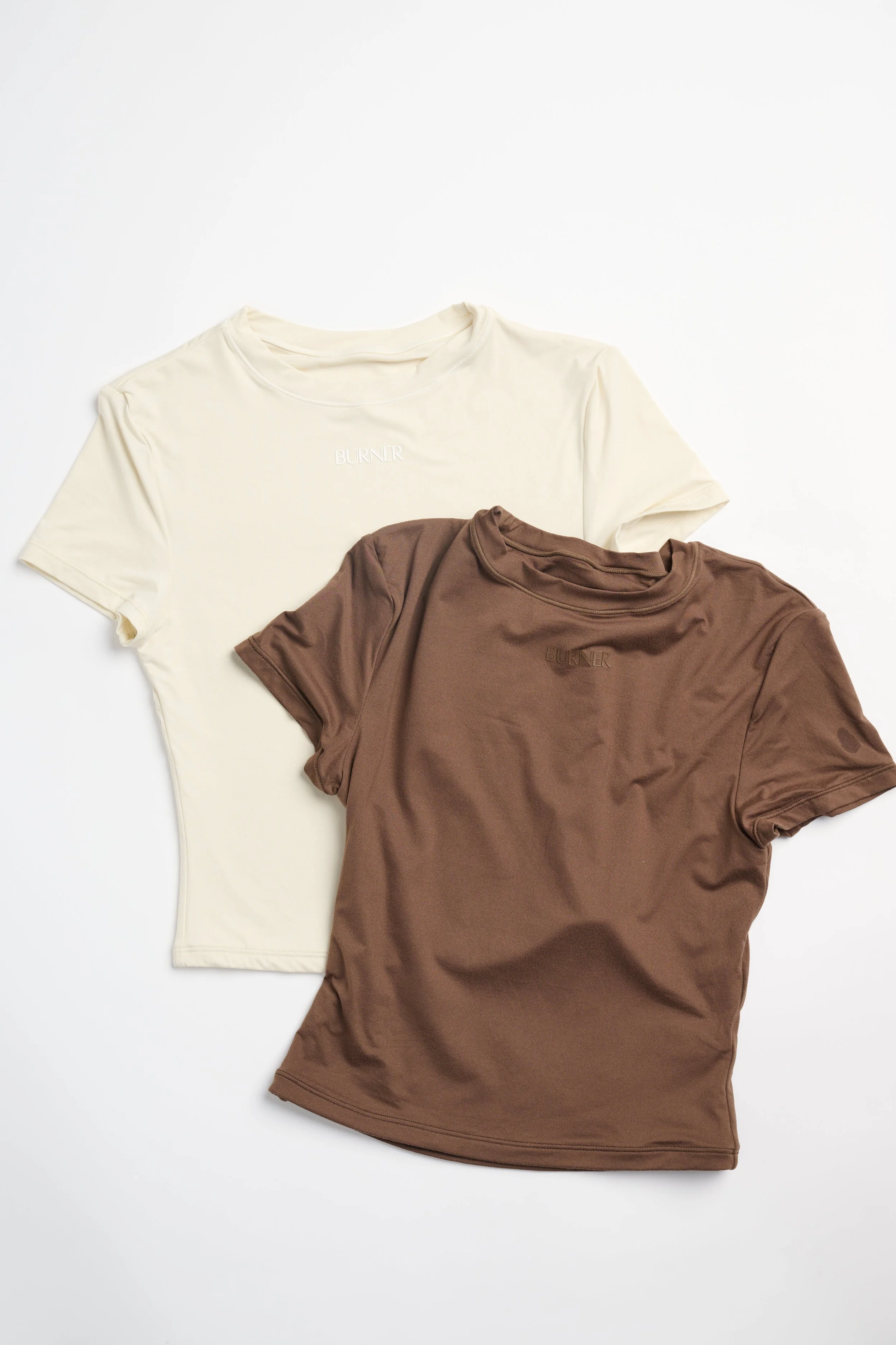 Two T-shirts, one cream and one brown, laid flat on a white background. The cream T-shirt has the word "BURNER" embroidered on the chest, while the brown T-shirt has the same word embroidered but less visible.