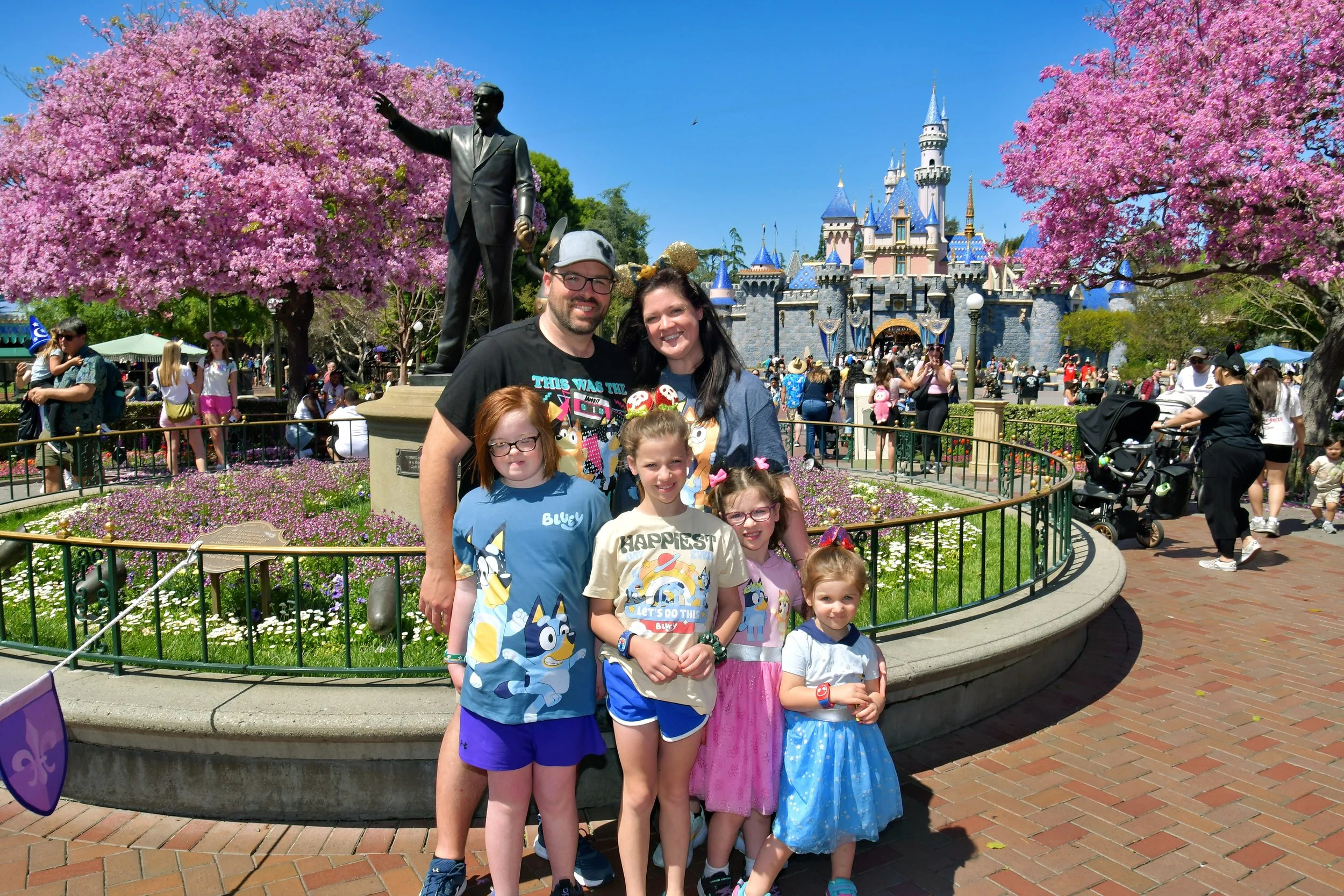 Family at Disneyland with Sleeping Beauty Castle in the background, children dressed in themed clothing, cherry blossom trees..