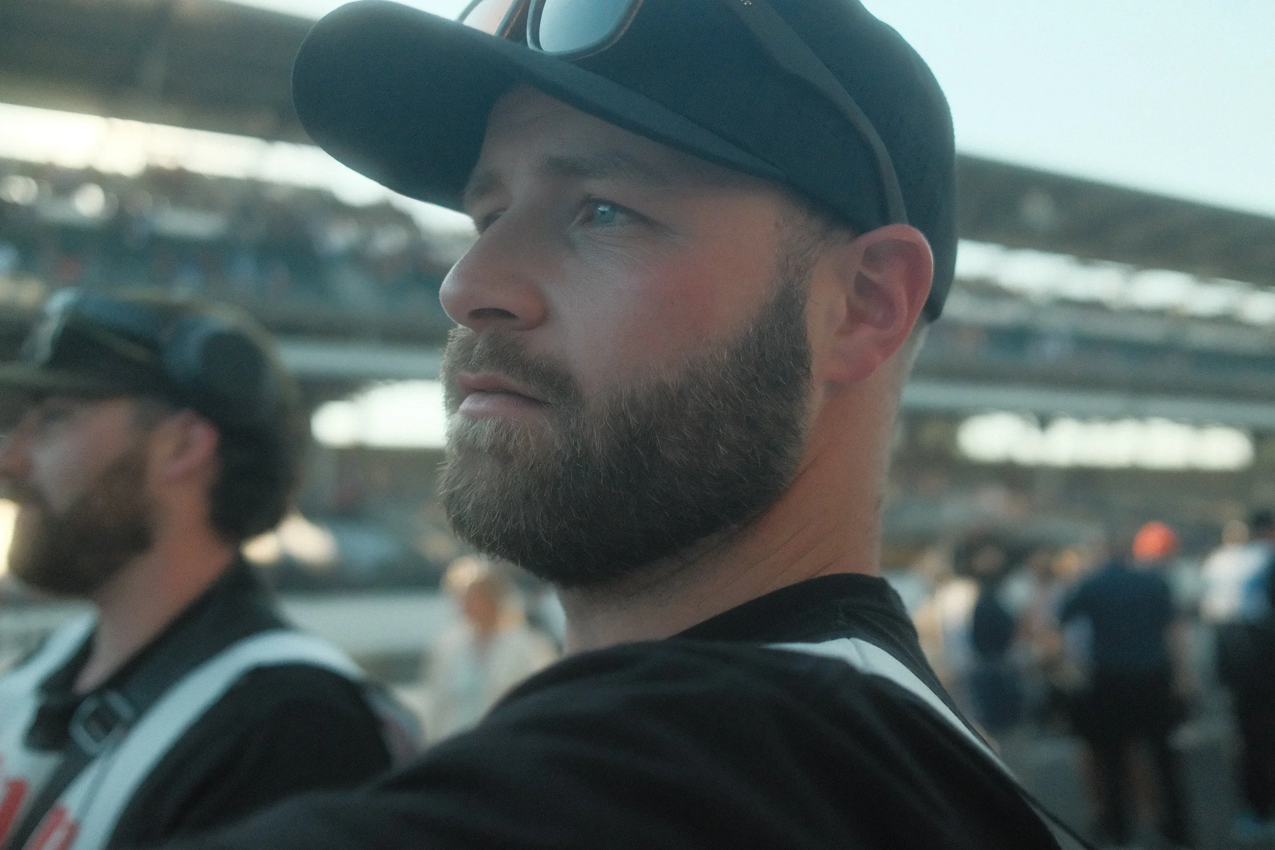 A man with a beard wearing a black cap and sunglasses on his head, looking to the side at an outdoor event or gathering.
