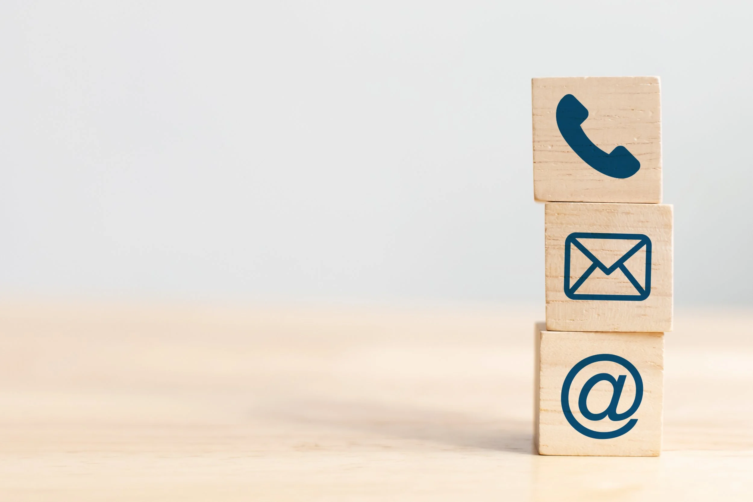 Three wooden blocks with communication icons: phone, email, and at symbol, stacked on a light wooden surface against a plain background.