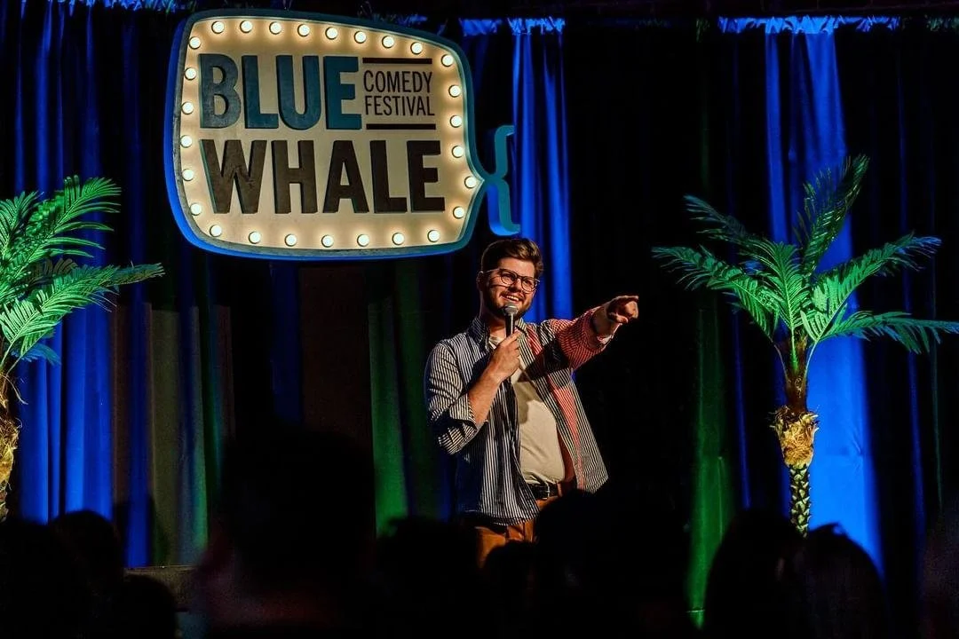 Colorado Springs comedian Russell Keller performing on stage holding a microphone, pointing, flanked by green palm trees, with a blue curtain background and a sign that says 'Blue Whale Comedy Festival'