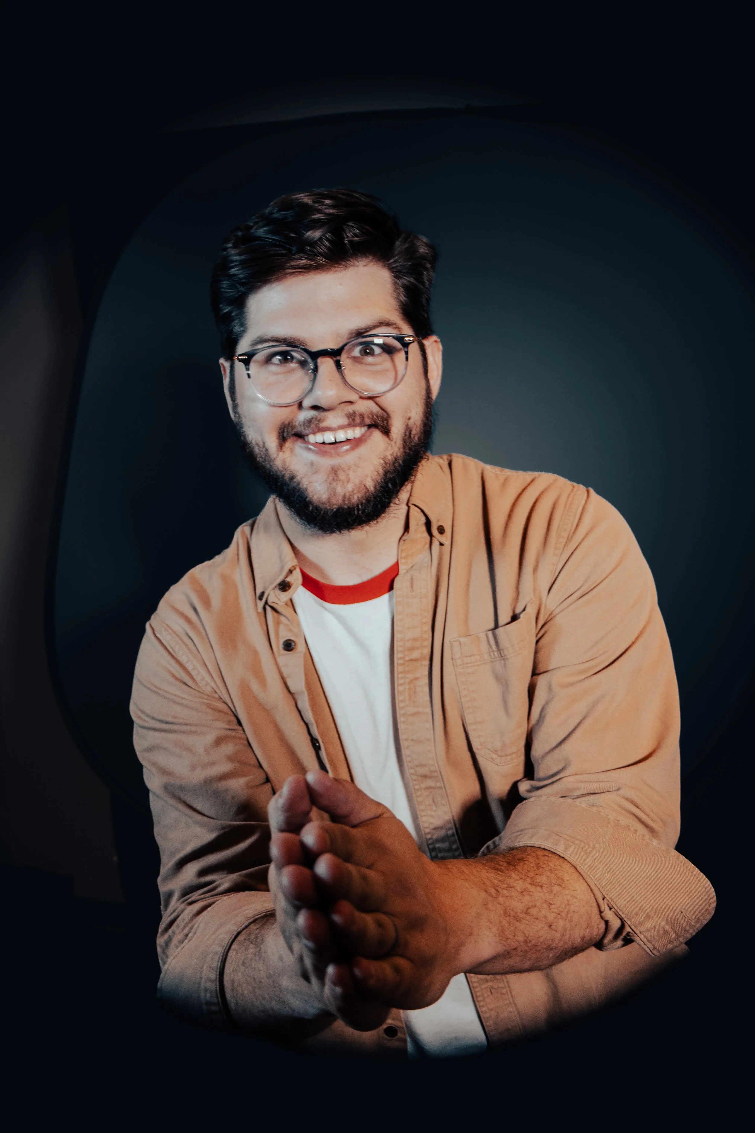 Colorado Springs comedian Russell Keller - a man with dark hair, glasses, and a beard smiling and looking at the camera, with a dark background, wearing a tan shirt over a white T-shirt.