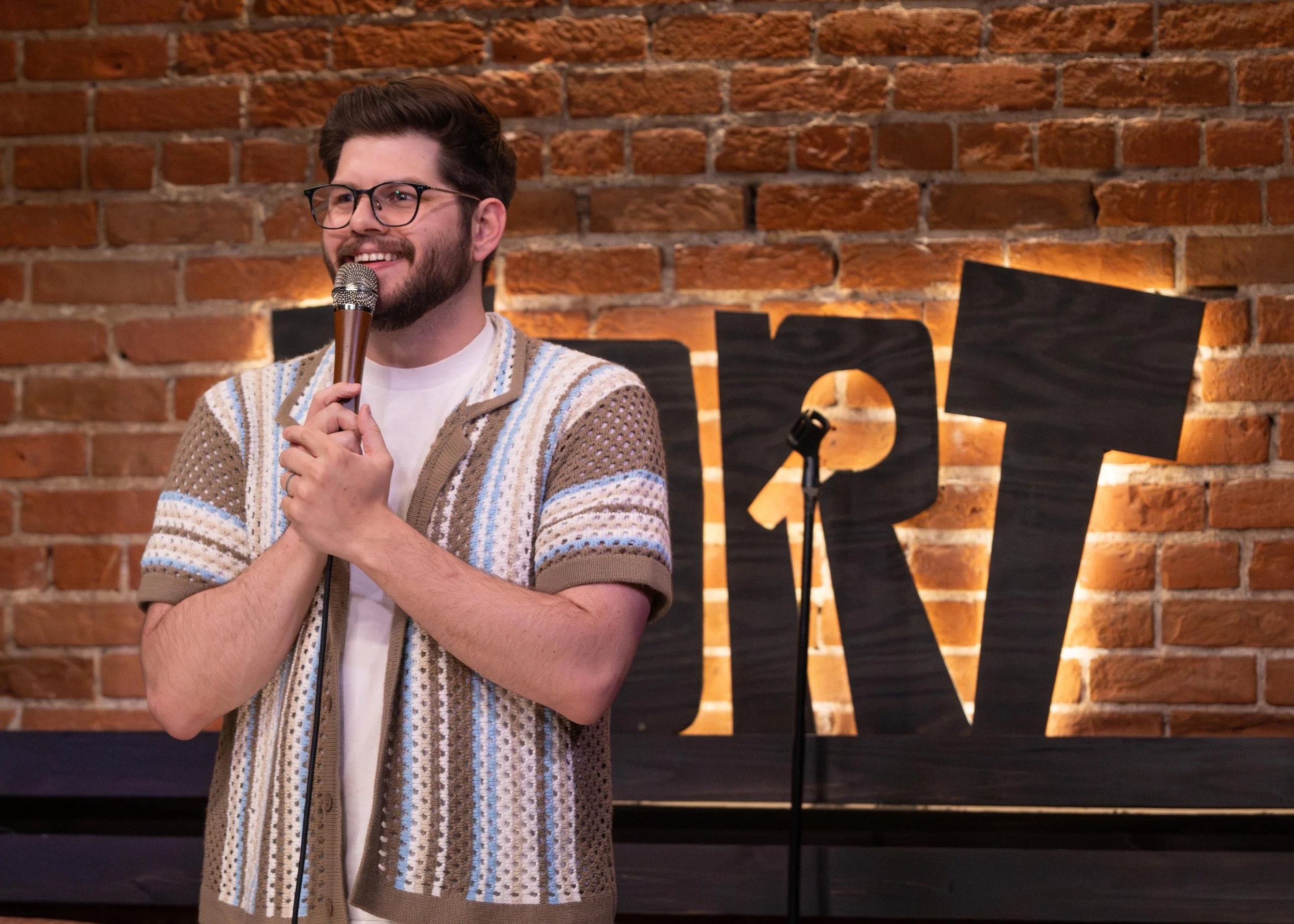Colorado Springs comedian Russell Keller, a man with glasses and a beard holding a microphone and smiling. He is wearing a patterned cardigan and standing in front of a brick wall with a black illuminated letters background, at the Comedy Fort