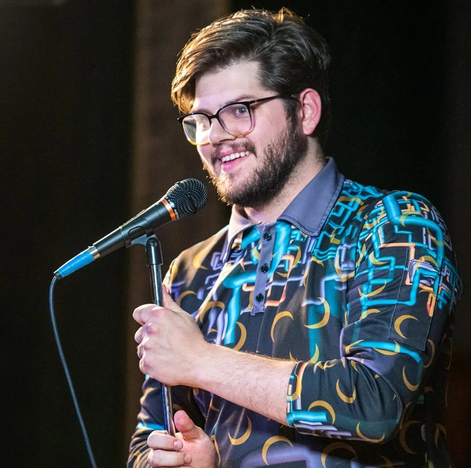 A male comedian with glasses, dark hair, and a beard holding a microphone and smiling, standing on a stage with a dark background. Russell Keller Colorado