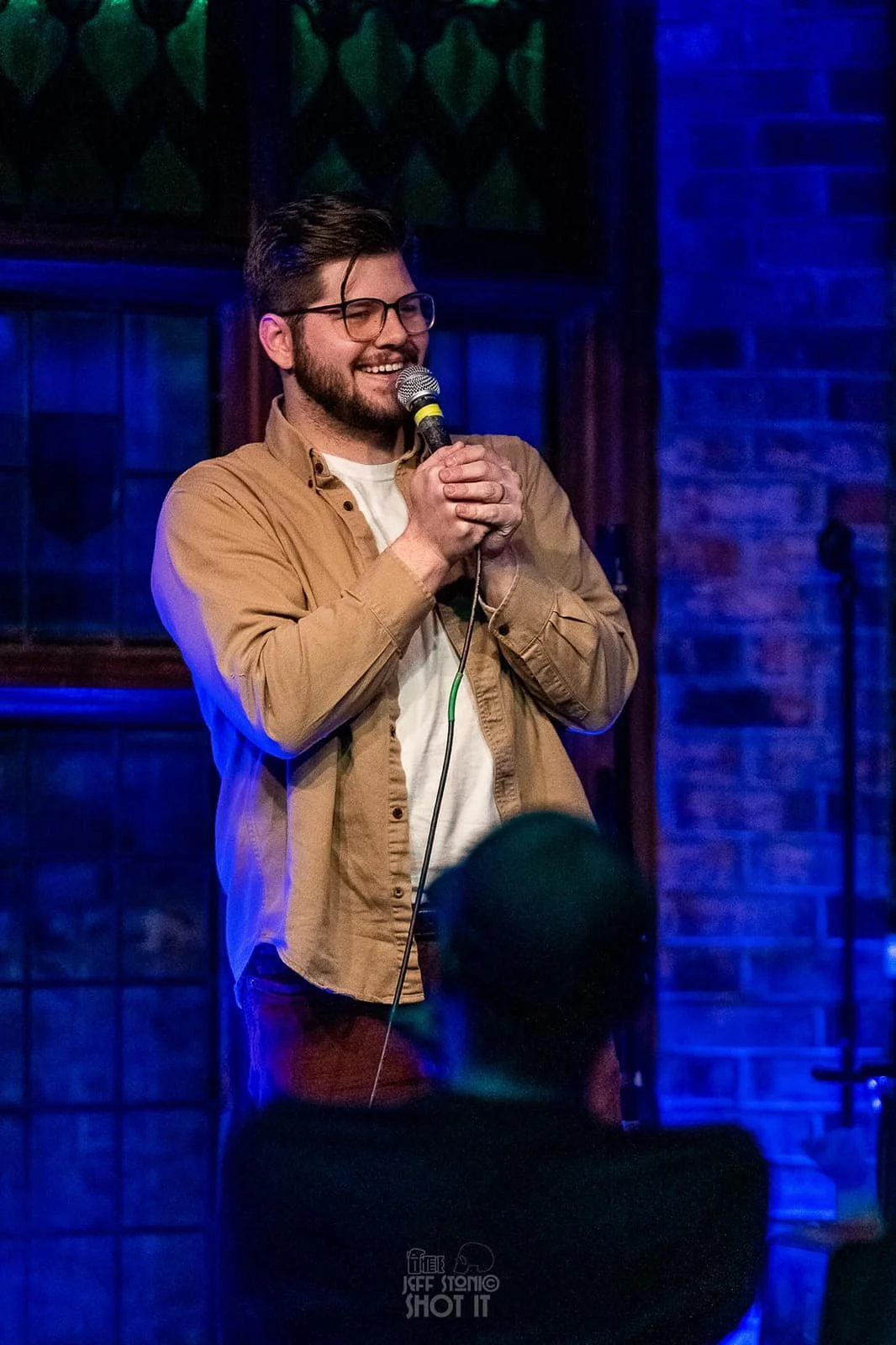 A male comedian with glasses and a beard, smiling, holding a microphone on stage at a comedy club or open mic night. He is wearing a tan jacket over a white shirt, with a brick wall in the background illuminated by blue lighting. Russell Keller Color