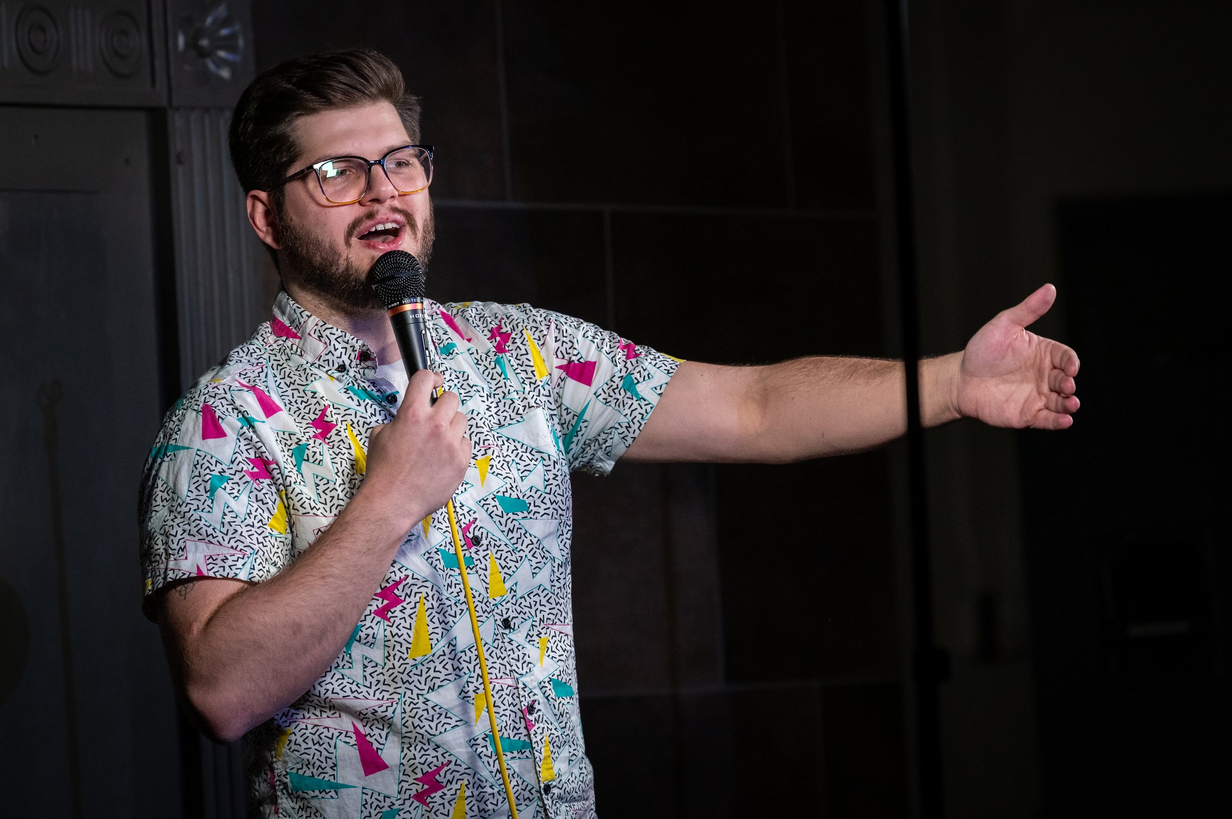 Colorado Springs comedian Russell Keller, a man with glasses and a beard, wearing a colorful patterned shirt, speaking into a microphone and gesturing with his right hand at the Pikes Punks Comedy Show.