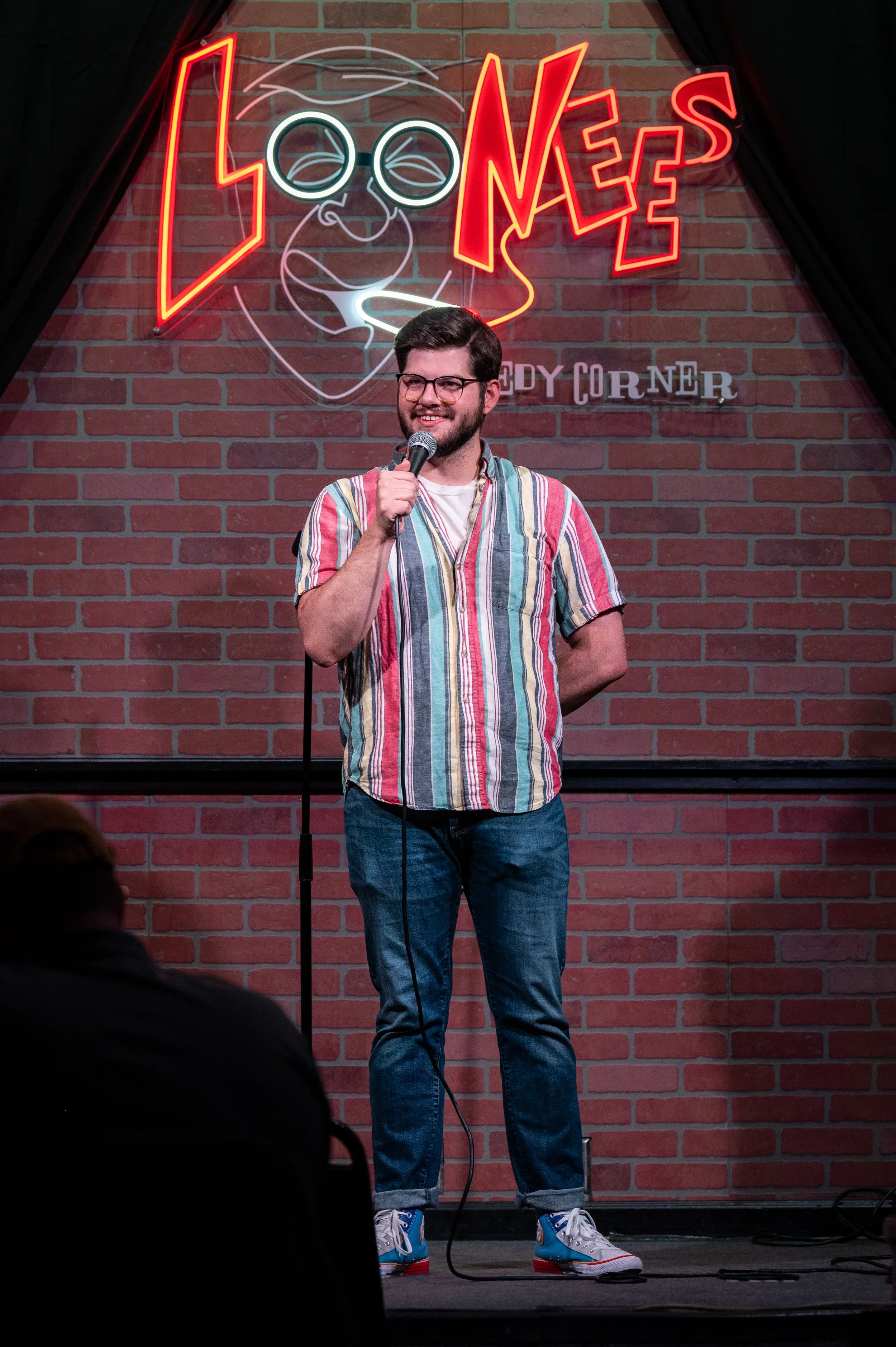 Colorado Springs comedian Russell Keler, is standing on stage at a comedy club. He is wearing a colorful striped short-sleeve shirt, jeans, and sneakers. The background features a brick wall with a neon sign that says LOONEES COMEDY CORNER