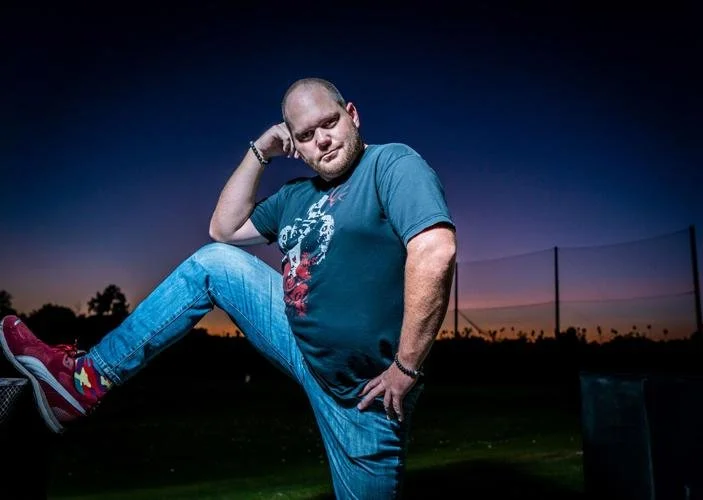 A man standing outdoors at sunset, striking a playful pose with one leg raised and one hand on his head, wearing a blue t-shirt, jeans, and red sneakers.