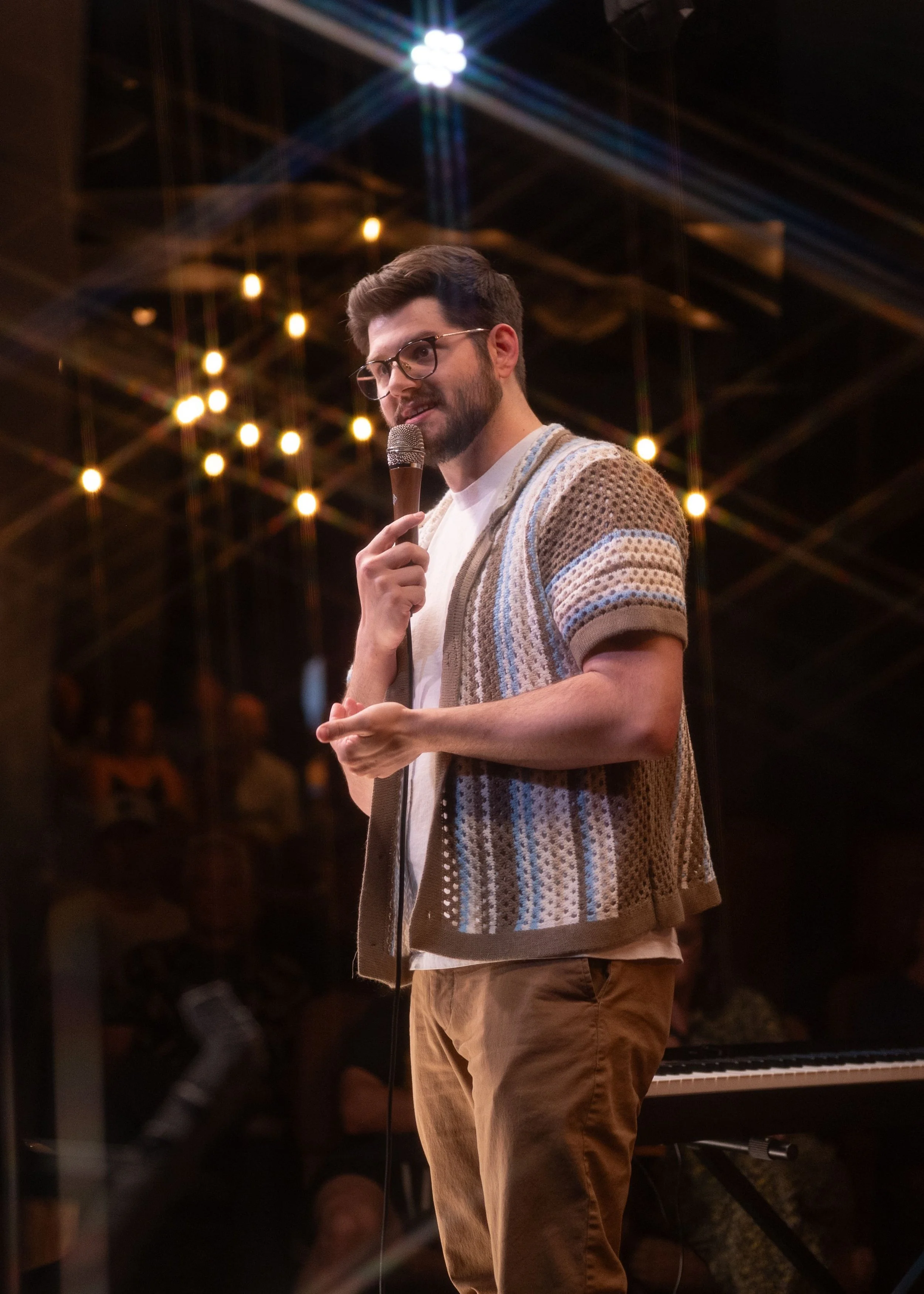 Colorado Springs comedian Russell Keller, a man with glasses and a beard holding a microphone, standing in front of a warmly lit background with string lights and a piano at The Comedy Fort