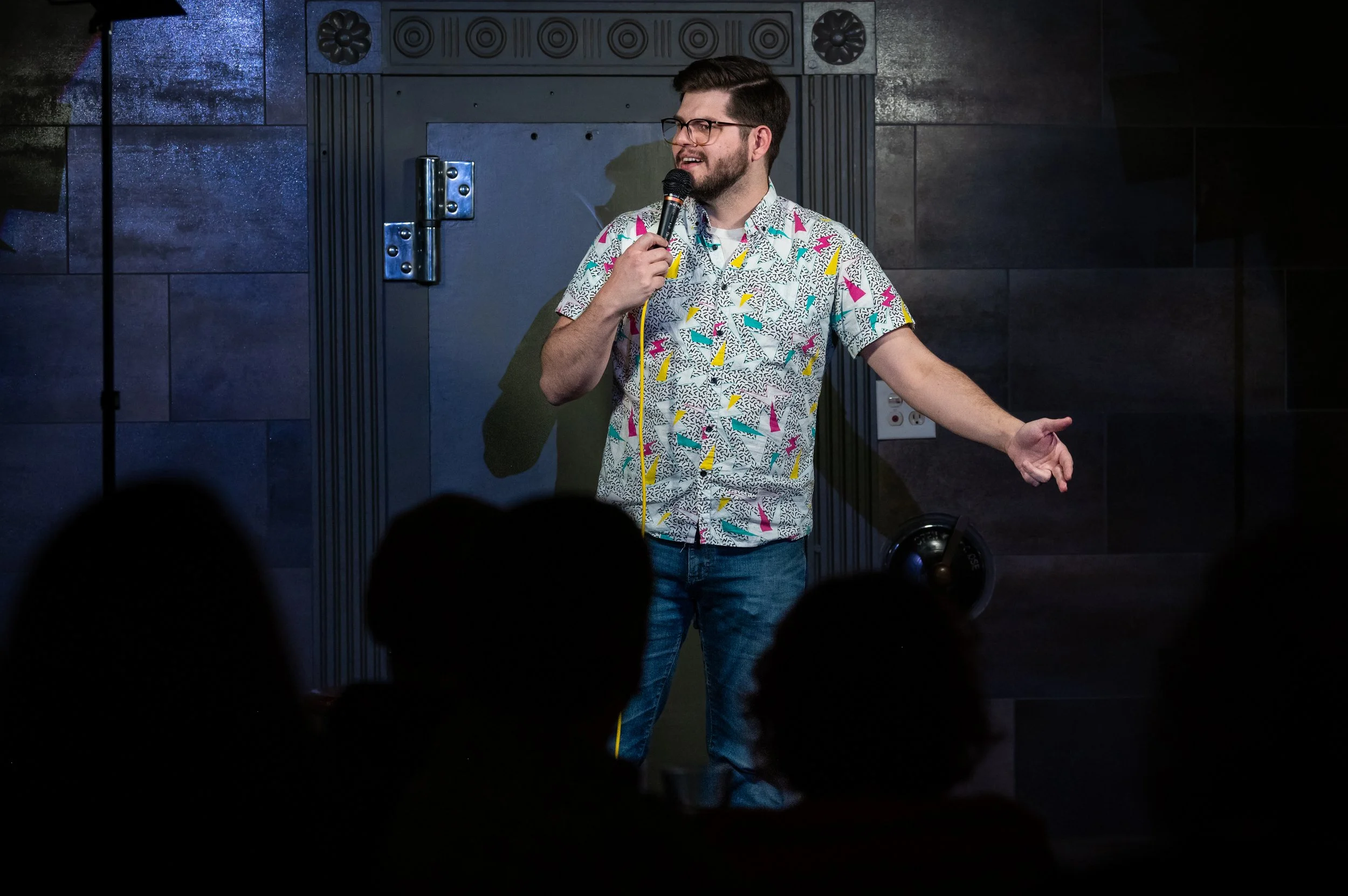 A male comic with glasses and a beard is performing stand-up comedy on stage, holding a microphone and gesturing with his left hand. He is wearing a colorful, patterned short-sleeve shirt and jeans. The background features a dark wall with a door, an