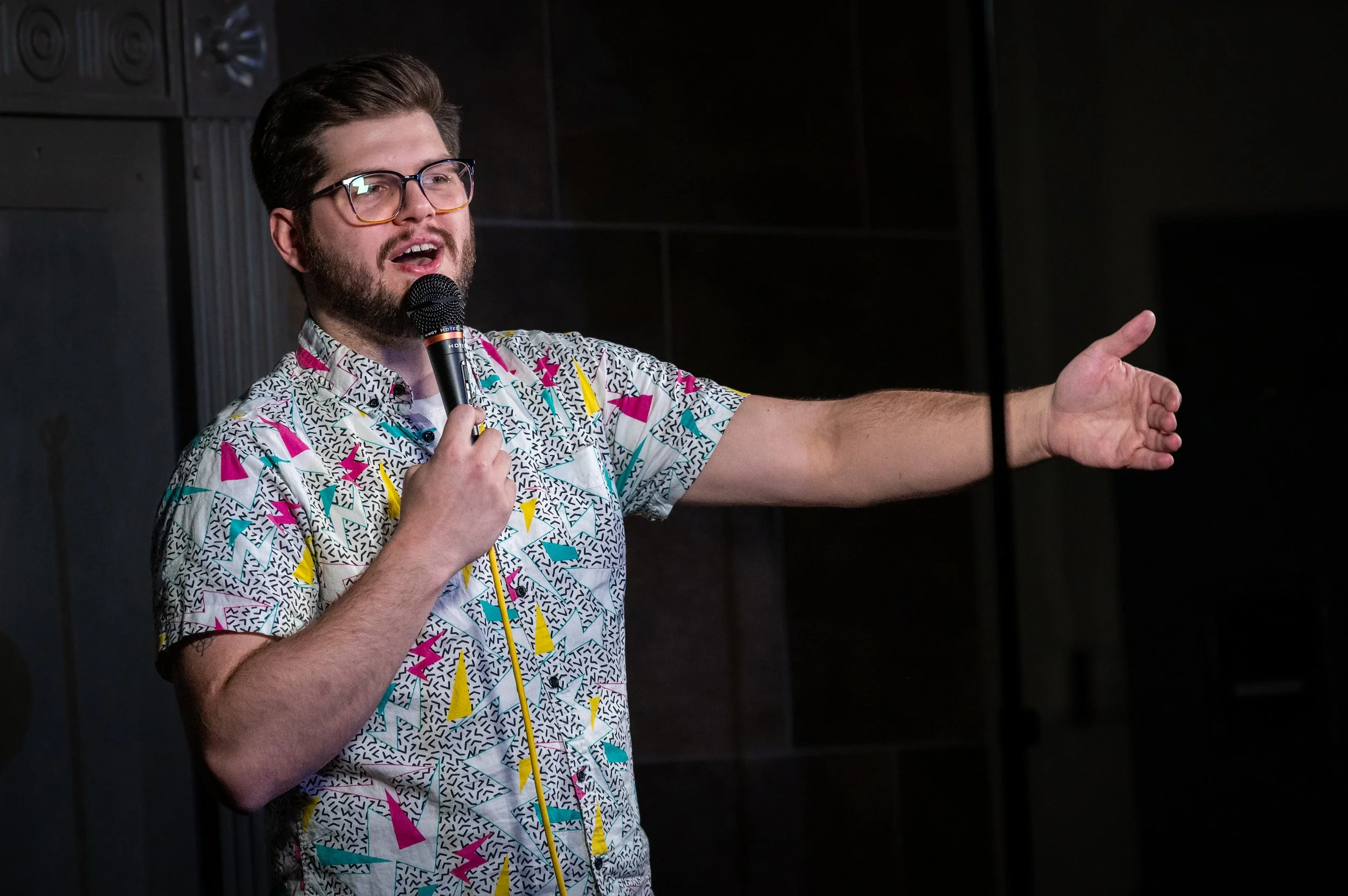 A man with glasses and a patterned shirt holding a microphone and gesturing with his right hand, speaking at an indoor comedy event. Russell Keller comedian colorado