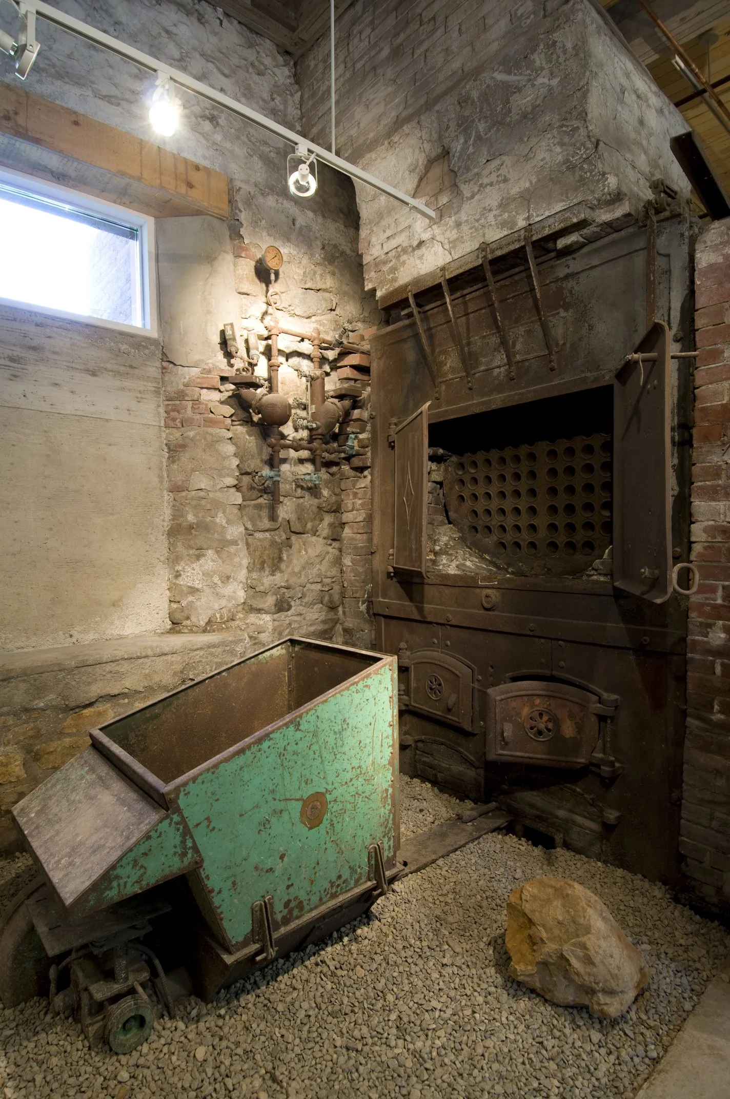 Indoor space with exposed brick and stone walls, featuring an old cast iron stove, a rusted green metal cart, and a large rock on a gravel floor. There is a small window and metal pipes on the wall.
