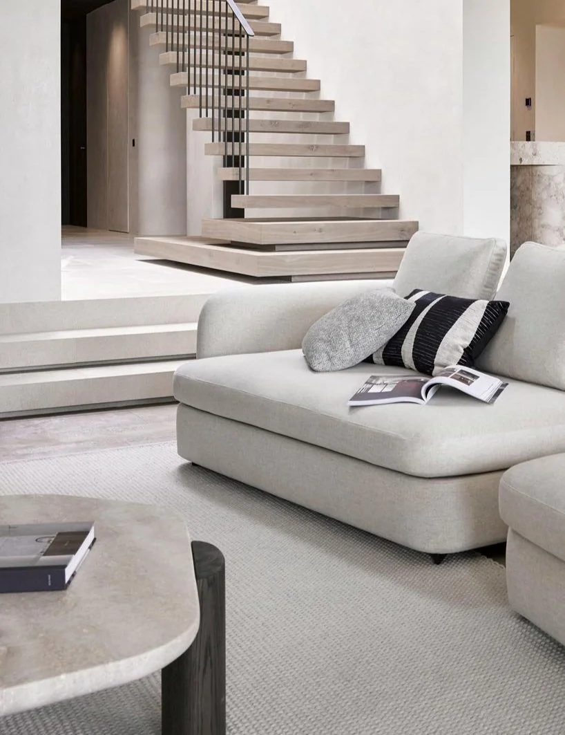 Interior of a modern living room featuring a white sofa with pillows, a marble coffee table, and wooden stairs with a metal railing in the background.