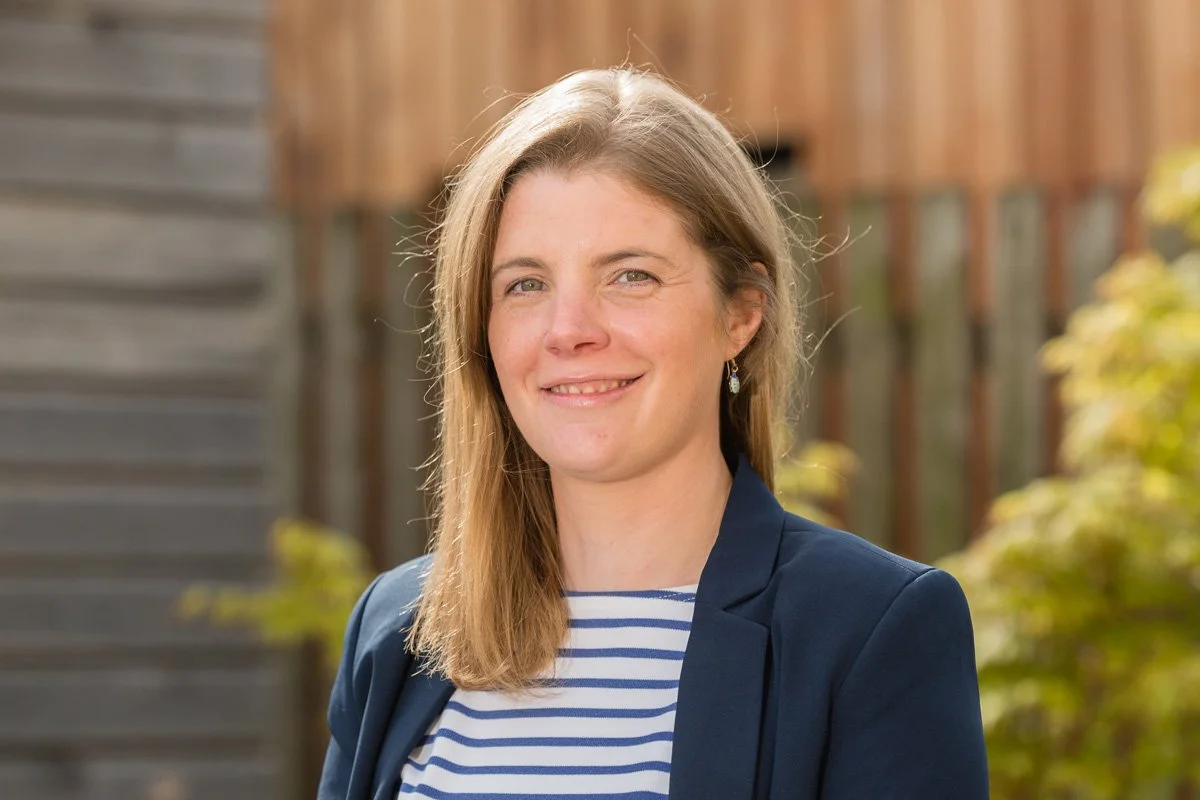 Photo of Steph Bridgeman, with long hair, wearing a blue blazer over a striped shirt, standing outdoors with a wooden fence and greenery in the background.