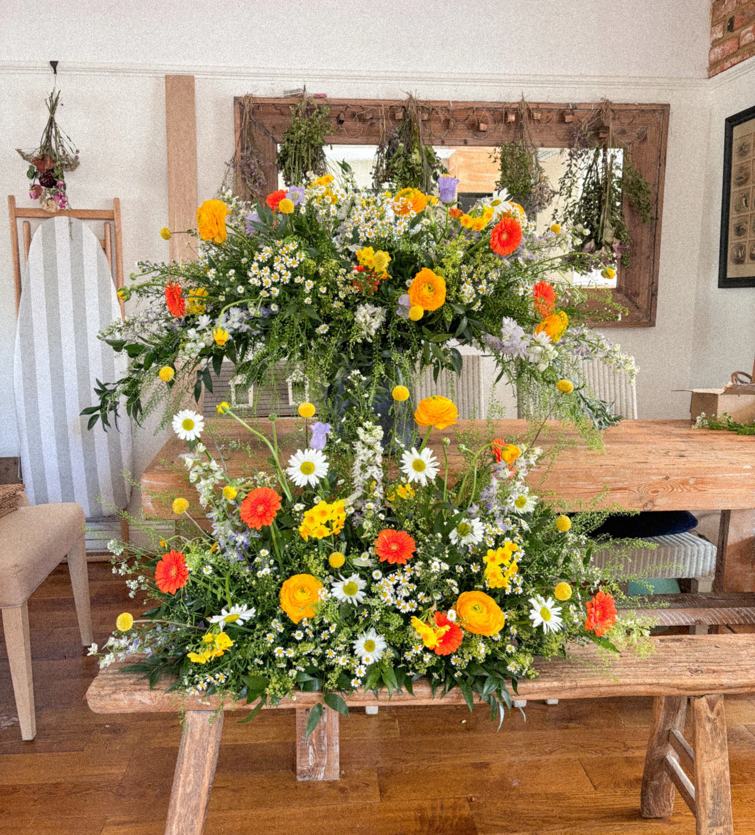 Two large floral arrangements with yellow, orange, white, and purple flowers on a wooden table inside a room.