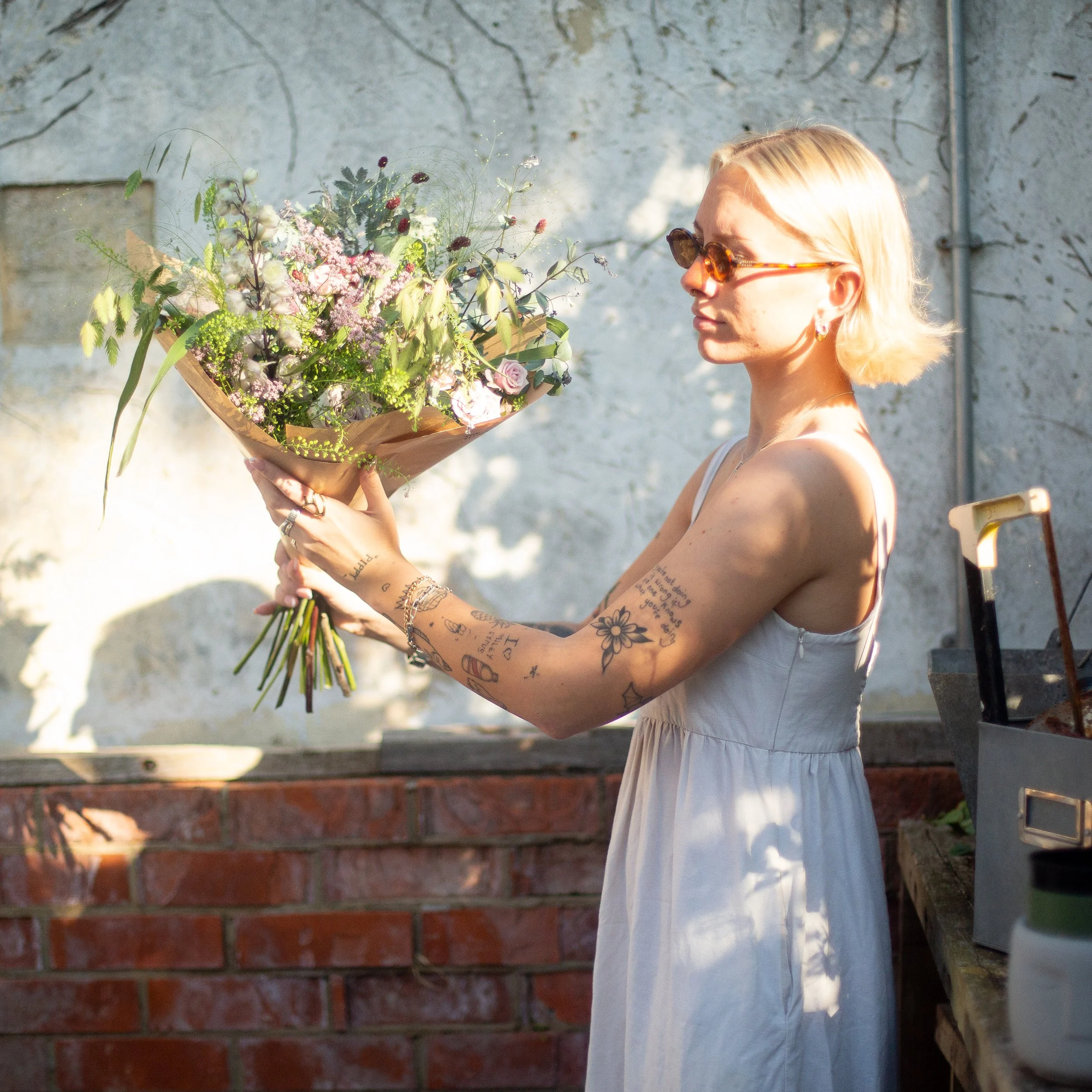 A woman with tattoos wearing sunglasses and a white dress holding a bouquet of pink, white, and purple flowers in front of a textured beige wall with sunlight casting shadows.