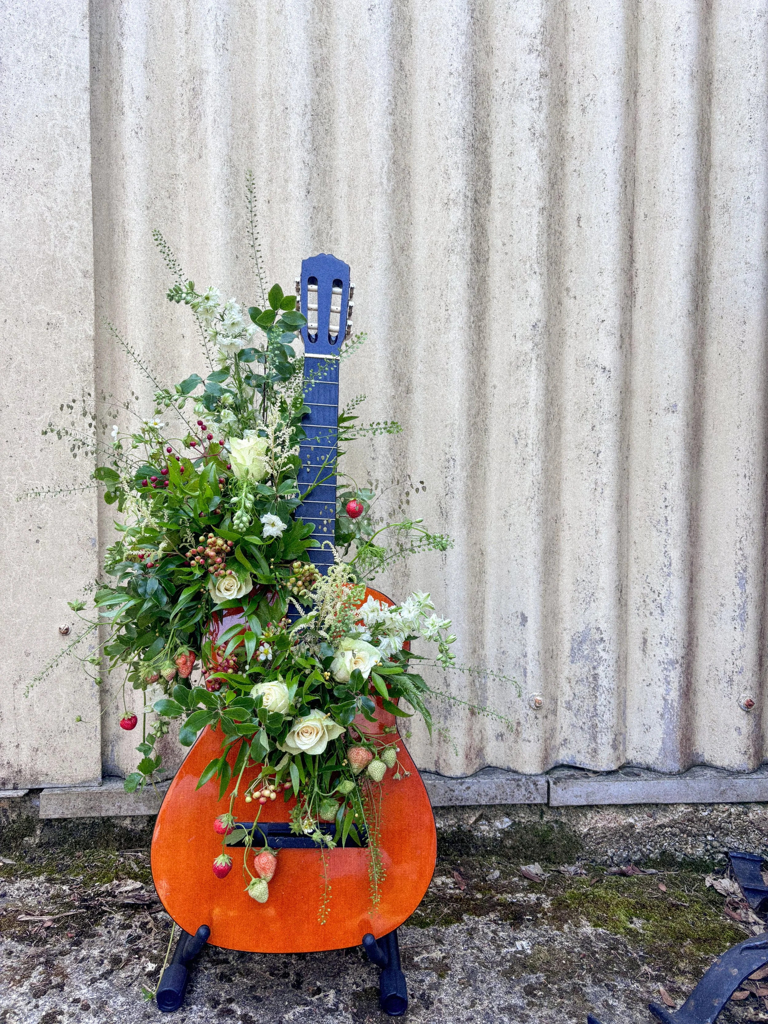 An orange guitar-shaped stand decorated with a bouquet of assorted white and green flowers and foliage, placed outdoors in front of a corrugated metal wall.