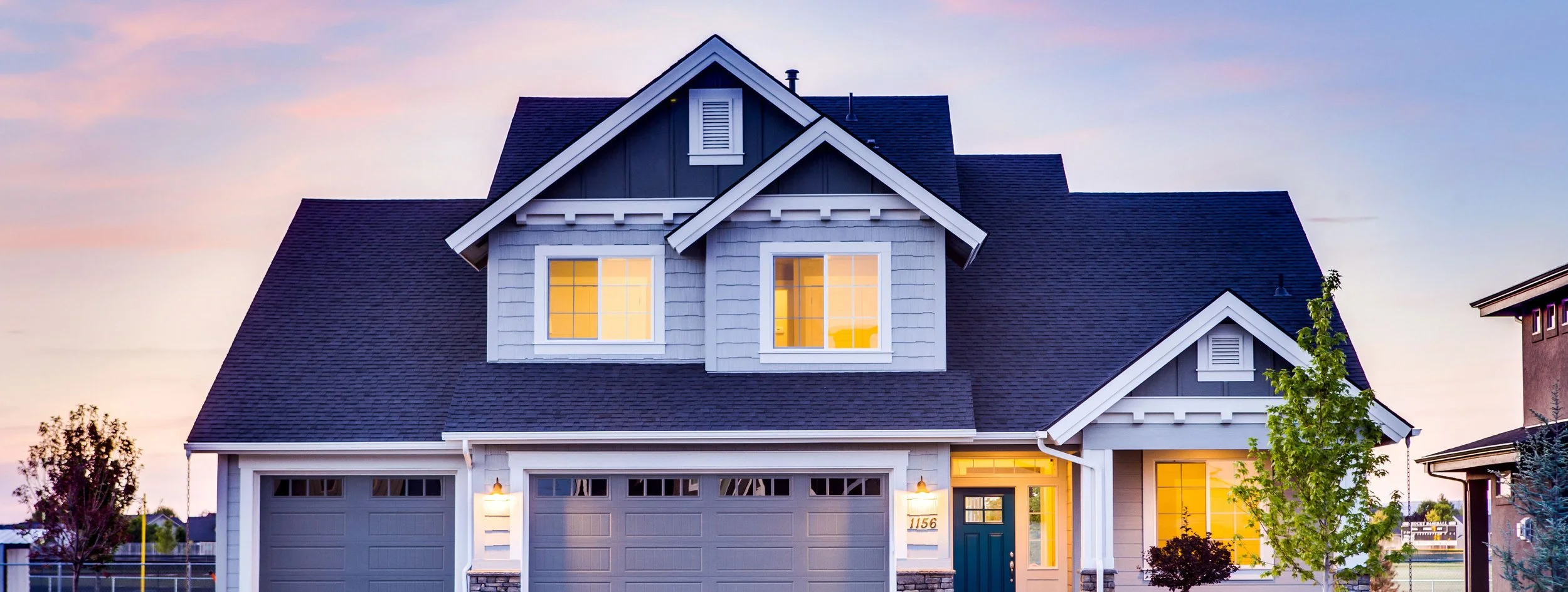 Exterior of a modern two-story house at dusk with warm yellow lights in its windows, gray siding, white trim, and a blue front door, with trees and neighboring houses nearby.