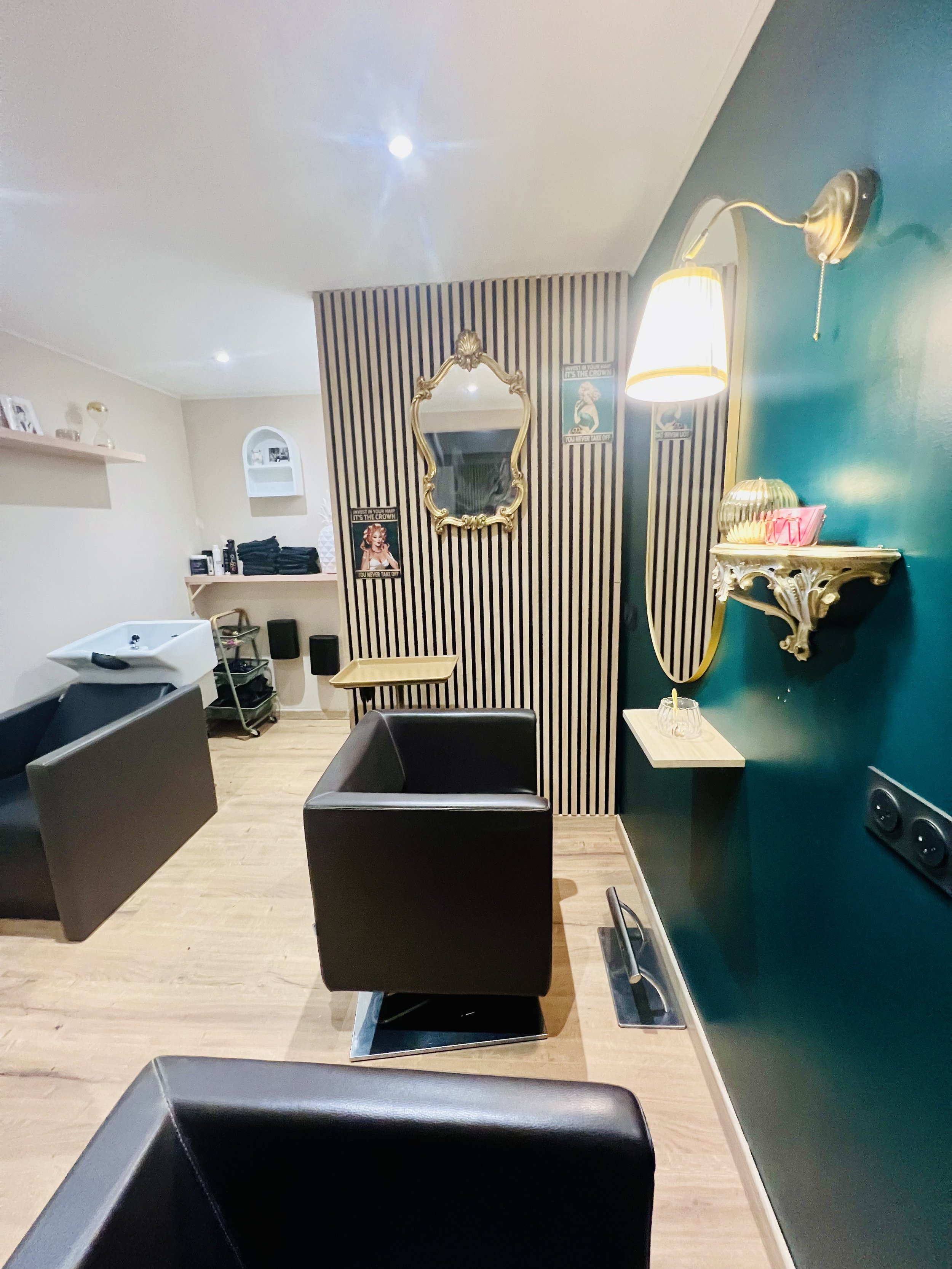 Salon de coiffure avec fauteuils en cuir noir, miroir doré, étagères ornées, et poste de lavage. Mur peint en vert avec lampes et décor vintage.