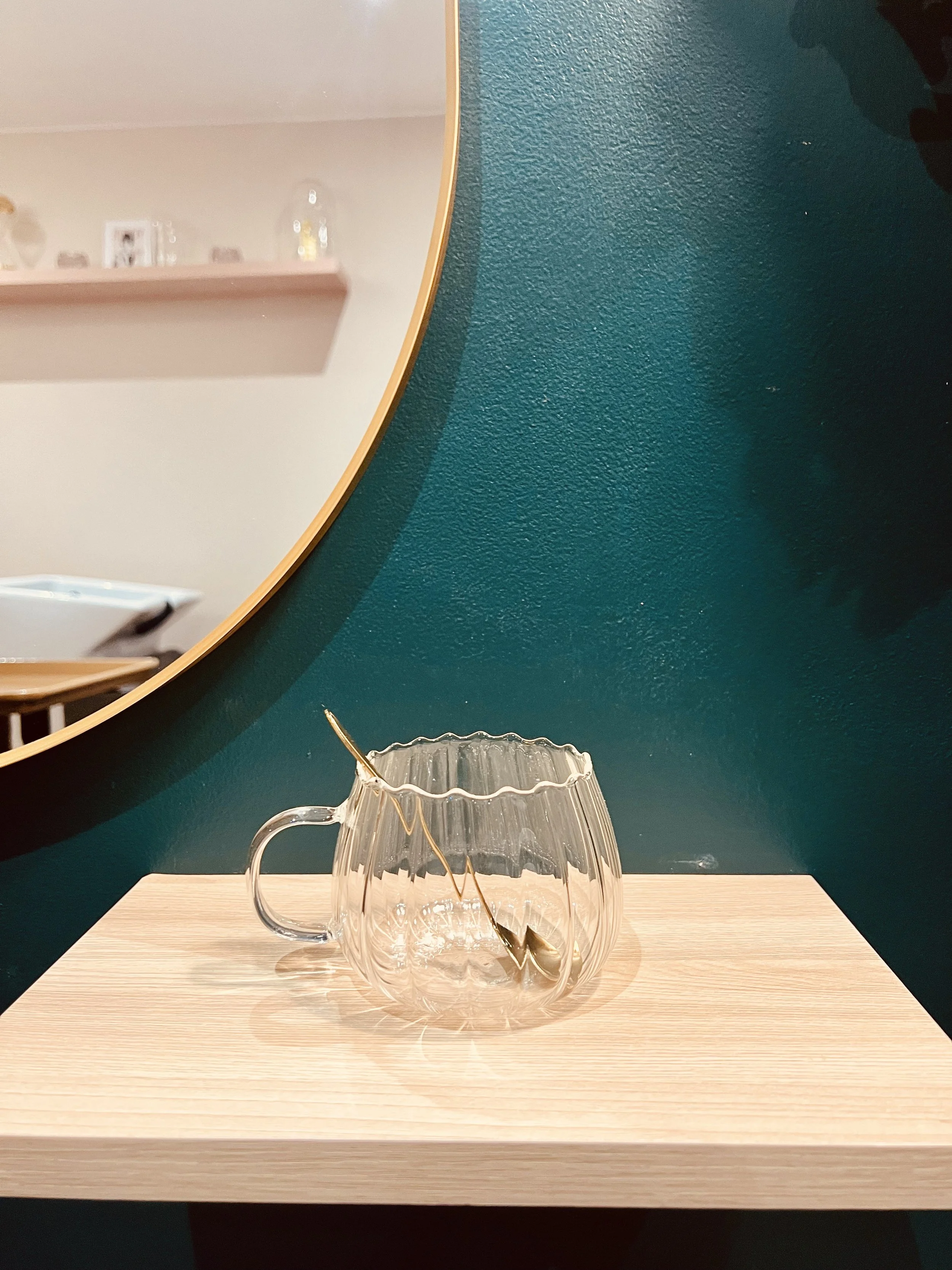 Une tasse en verre transparent avec une poignée, posée sur une table en bois, devant un mur vert foncé et un miroir rond.