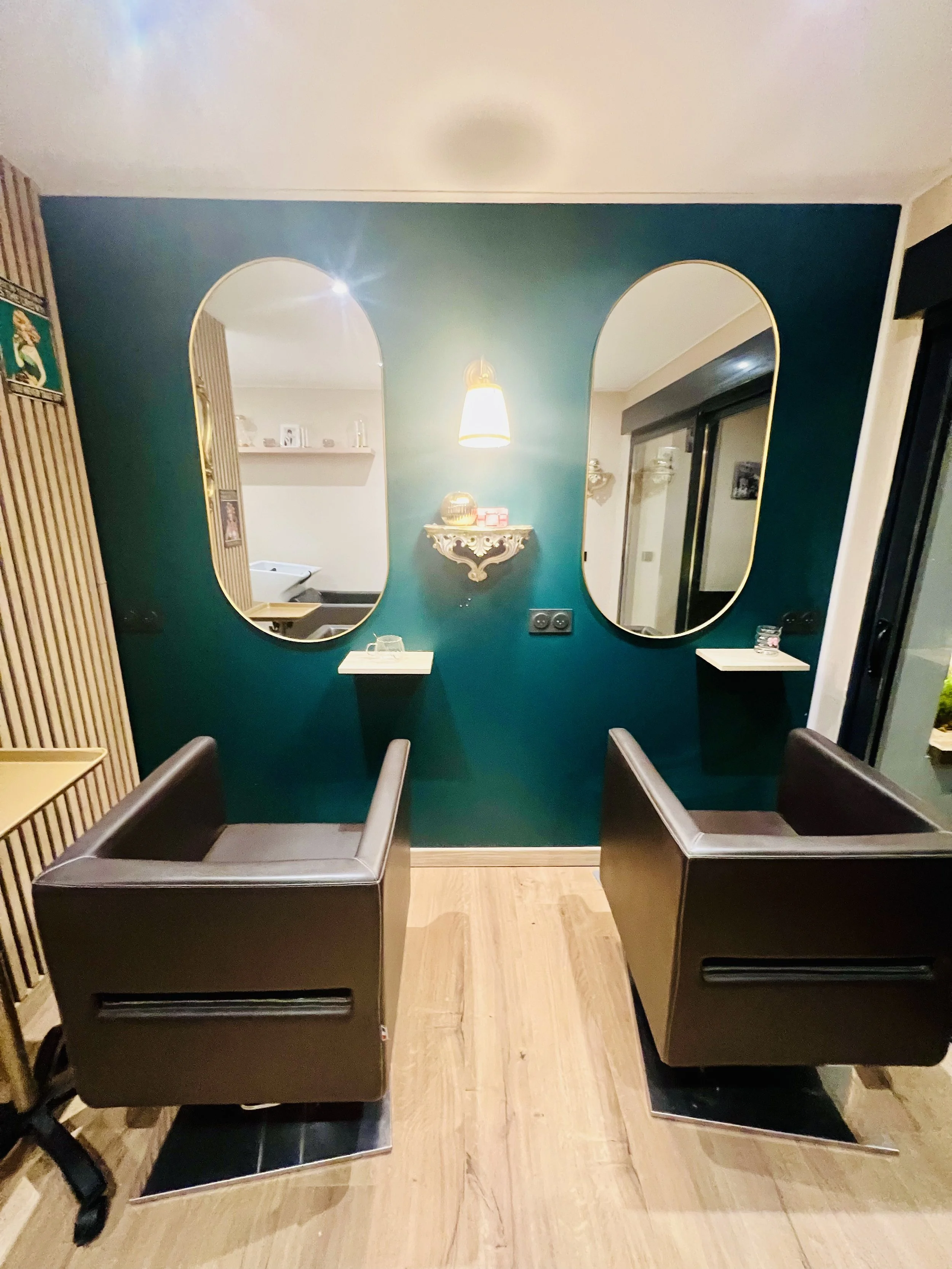 Salle de coiffure avec deux fauteuils en face de deux grands miroirs ovales sur un mur vert foncé.
