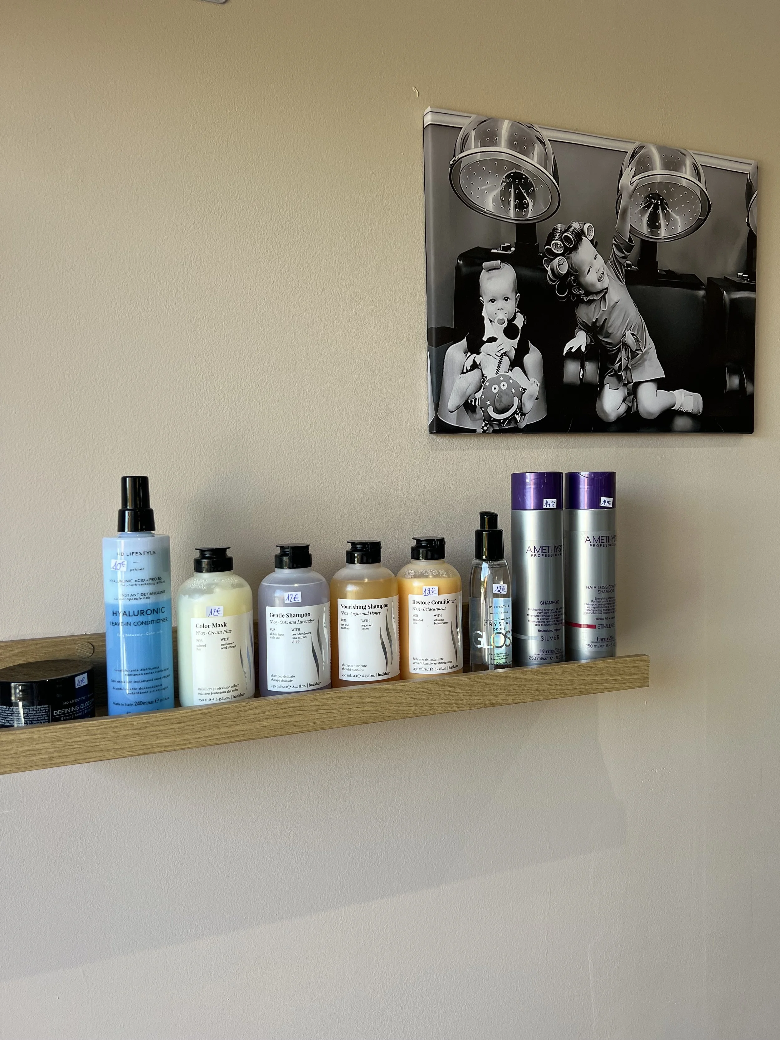 Prise en photo de plusieurs produits capillaires sur une étagère en bois, avec un tableau noir et blanc d'enfants dans un salon de coiffure en arrière-plan.