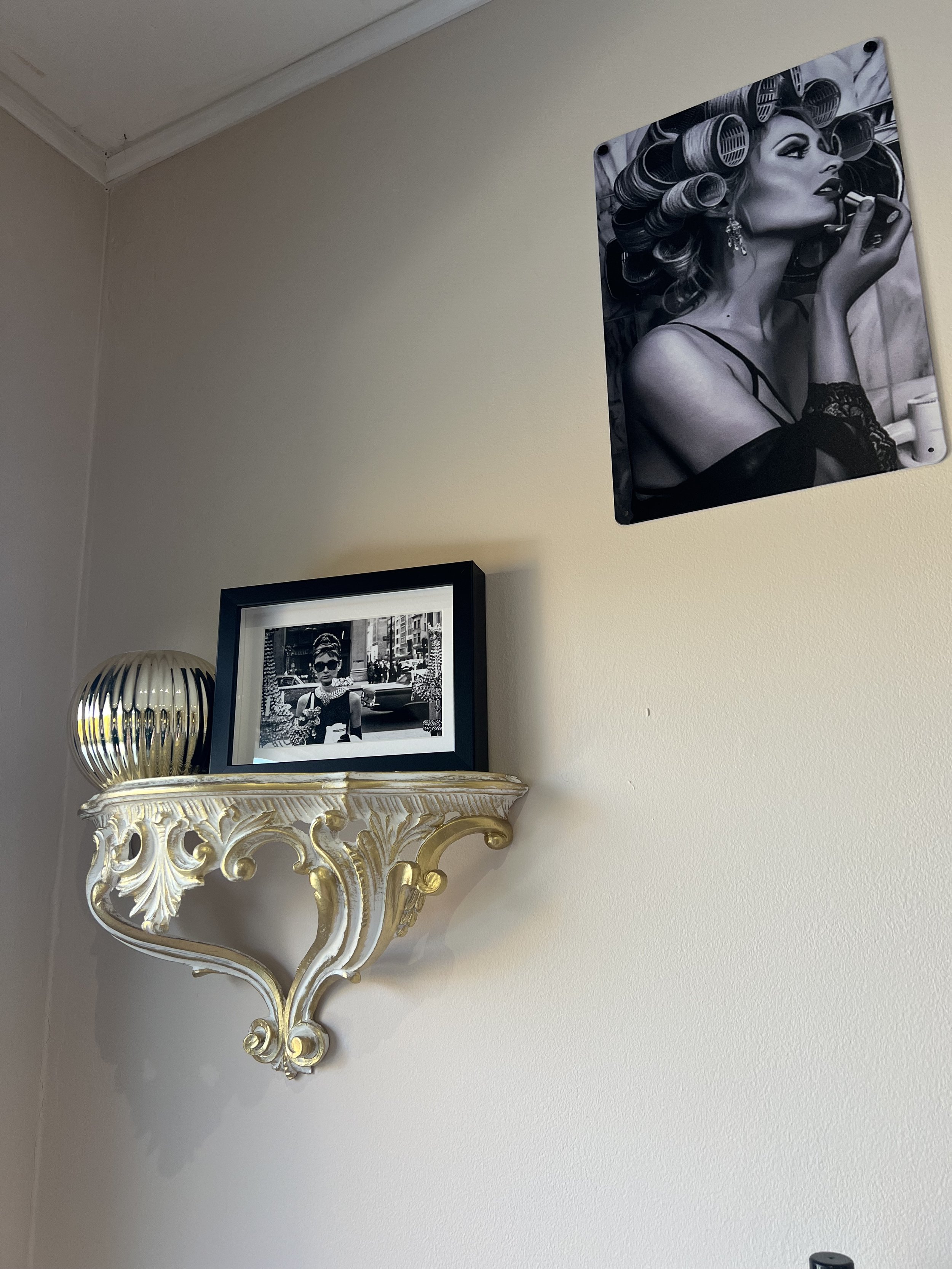 Décor de mur avec une photo en noir et blanc d'une femme avec des rouleaux de coiffure dans les cheveux, une petite étagère dorée avec un cadre photo en noir et blanc d'une femme portant des lunettes de soleil, et une décoration ronde en métal argent