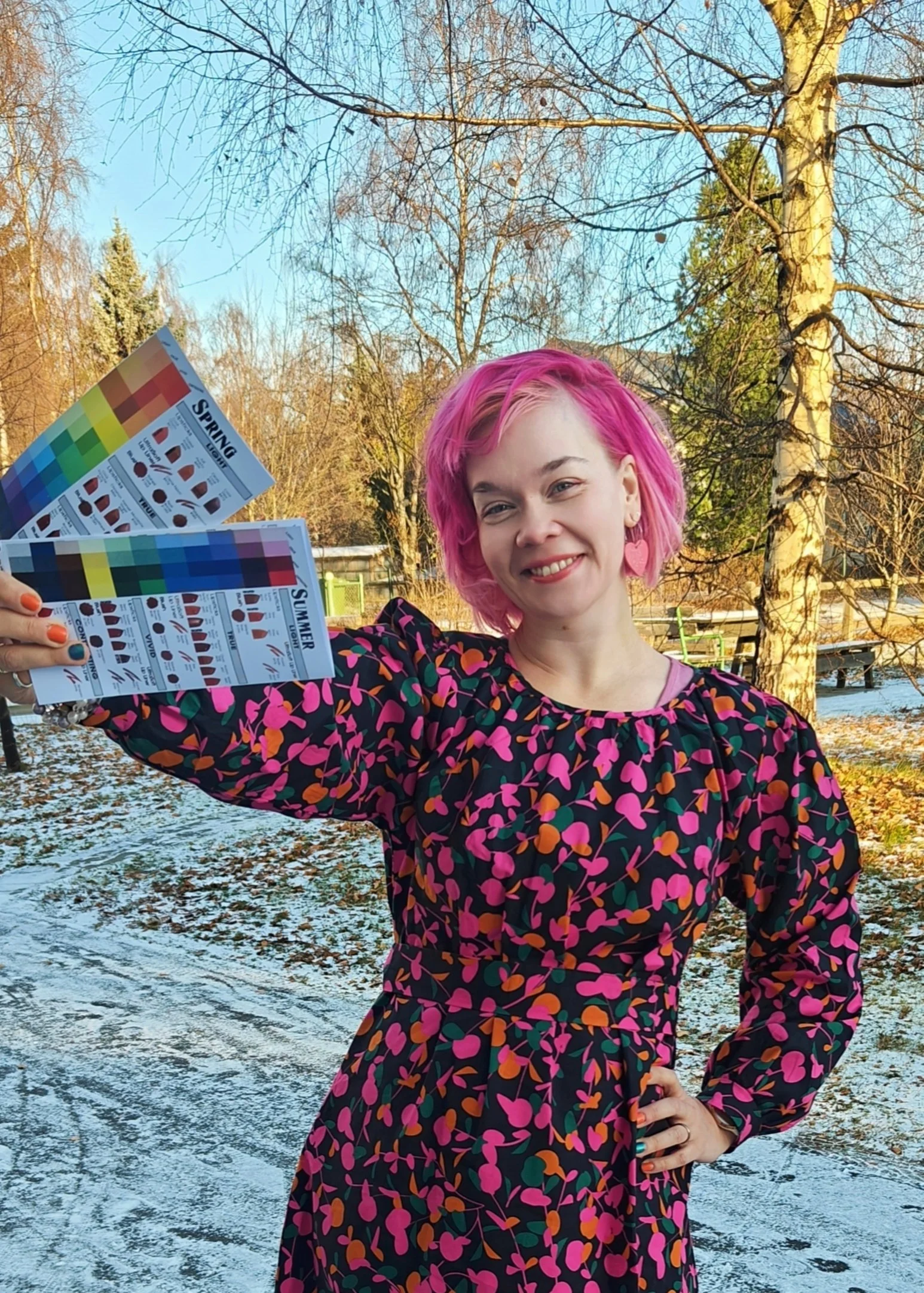 Nainen, jolla on pinkit hiukset pitelee värikarttoja ulkona lumisessa puistossa.