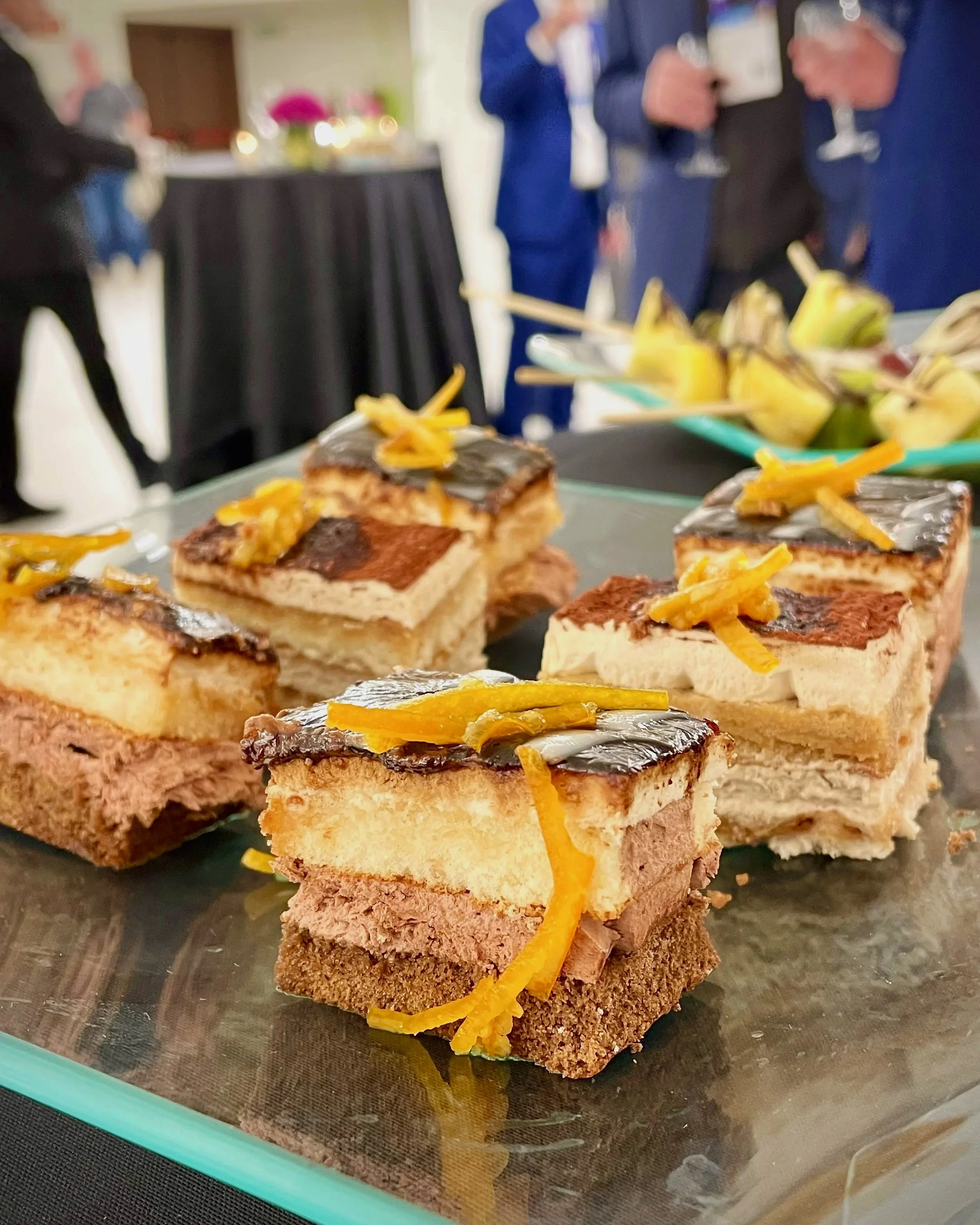 Pieces of layered cake with frosting and orange zest on top, displayed on a glass serving tray at a social event.