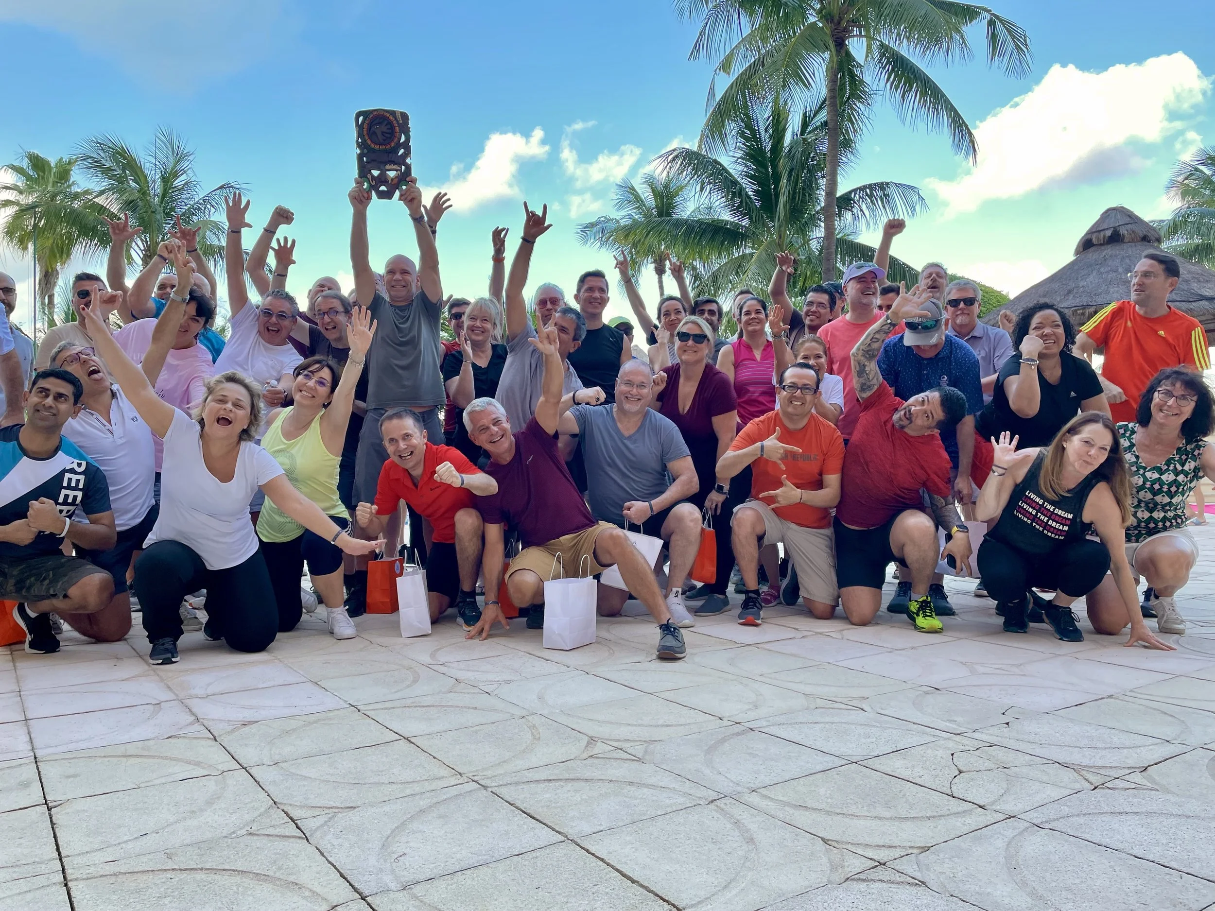 Celebratory group photo from a Sales Kickoff team-building event—employees on a tropical beach raising a trophy after a friendly competition, highlighting internal engagement strategy.