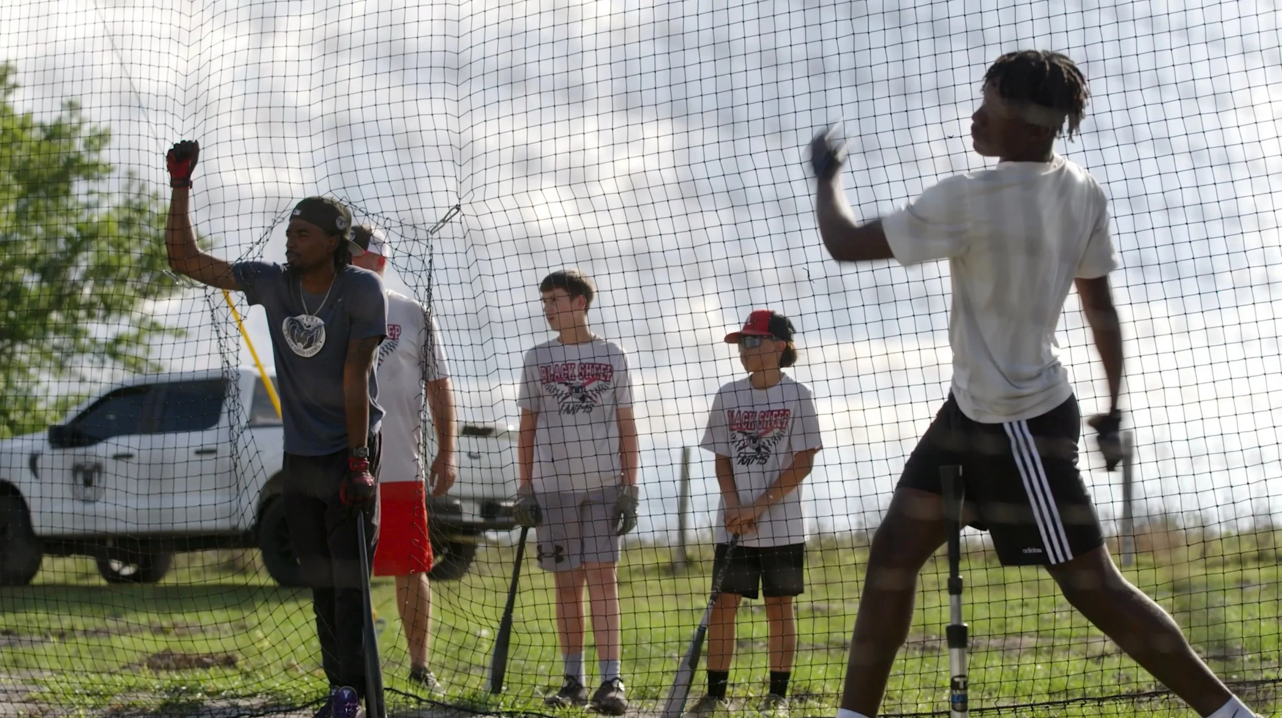 A group of boys practicing baseball inside a batting cage on a grassy field, with a white truck and trees in the background.