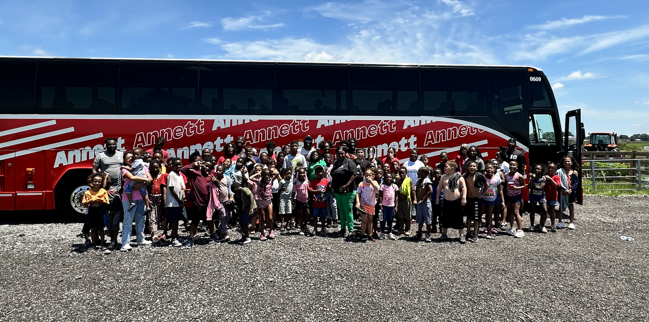 Group of children and adults standing in front of a large red bus with the name 'Annett' written on it, on a gravel lot under a blue sky.