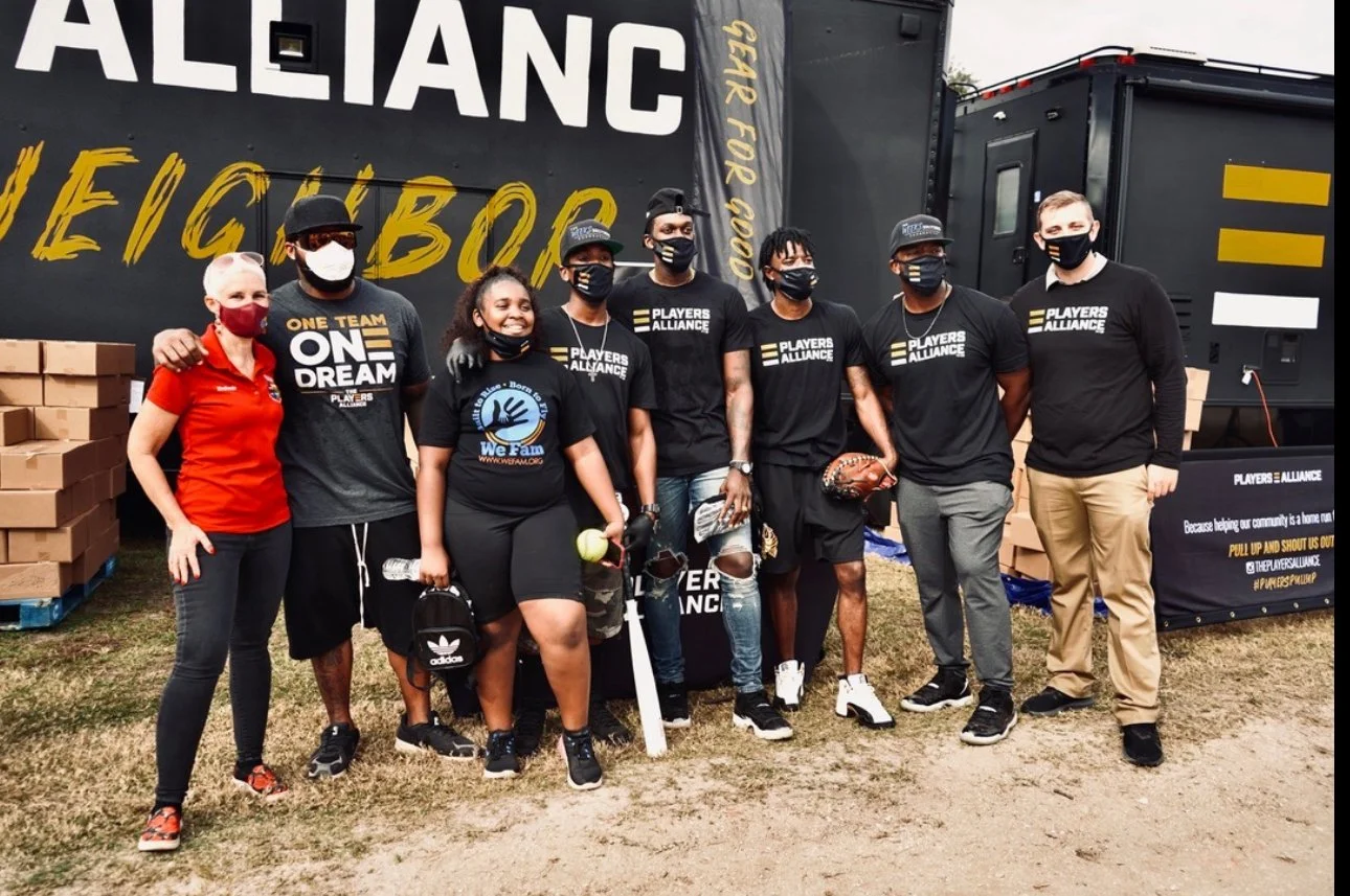 Group of nine people standing outdoors, wearing masks, in front of a black tent with yellow and white text, holding sports equipment, some with arms around each other.