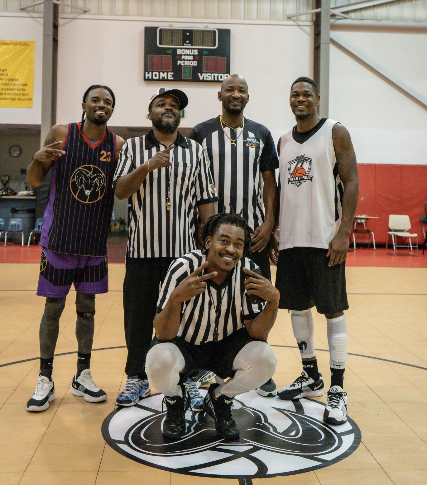 Five basketball players and referees posing on a court, with four standing and one squatting in front. The players are in uniforms with bright colors and logos, and the referees are in black-and-white striped shirts. They are smiling and making gestures, with a scoreboard in the background.