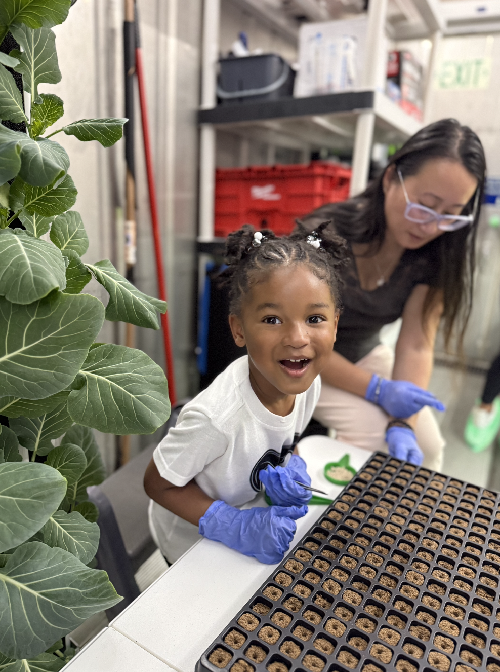A young girl with braided hair and a white t-shirt is smiling excitedly while working with a woman in the background. They are planting seeds in small pots, wearing blue gloves, inside a workshop or classroom setting with plants and tools around.