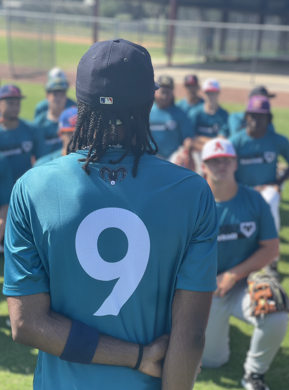Back of a baseball player in a blue uniform with the number 9 speaking to a team during a practice or game, with teammates in the background on the field.
